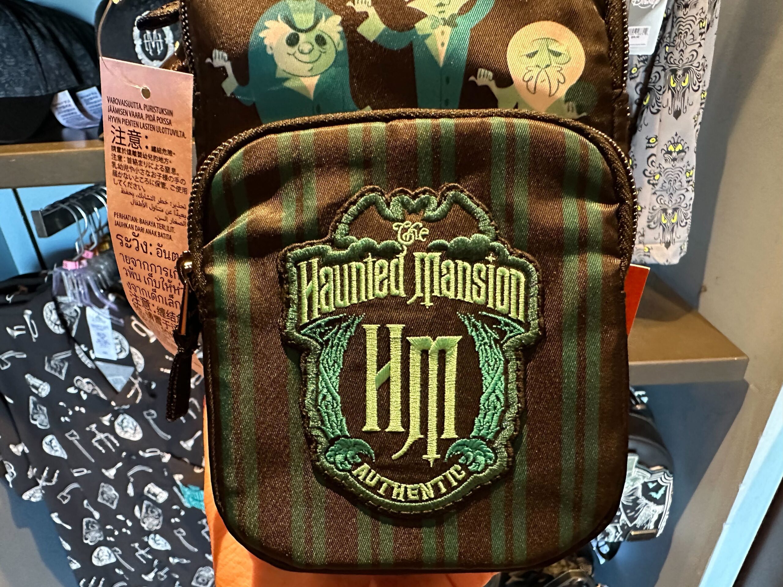 A small brown shoulder bag with green stripes, featuring "The Haunted Mansion" logo on the front, displayed in a store.