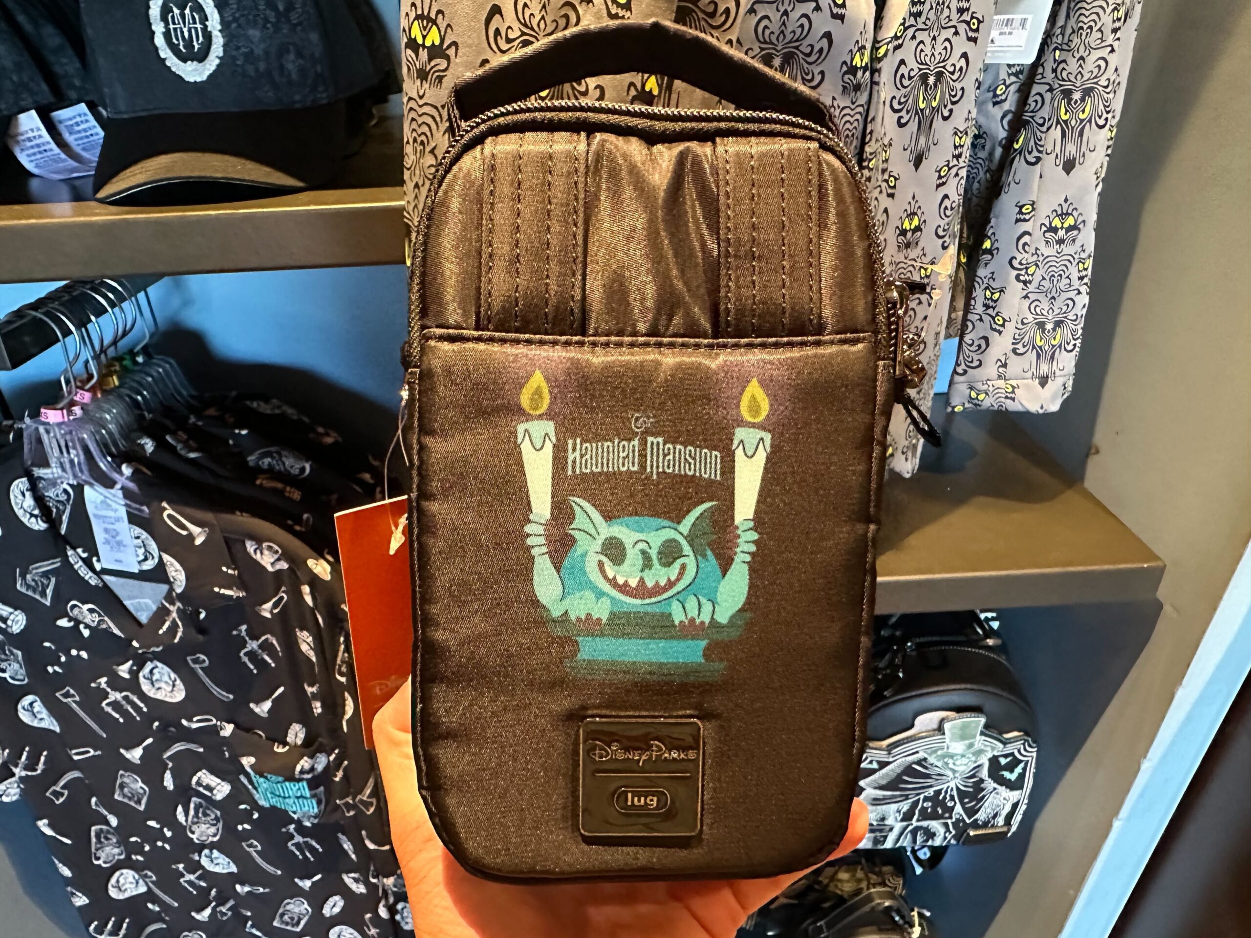 Close-up of a Haunted Mansion-themed crossbody bag with an embroidered ghost and candles design. Disney Parks tag visible at the bottom. Displayed on a shelf with other themed merchandise.