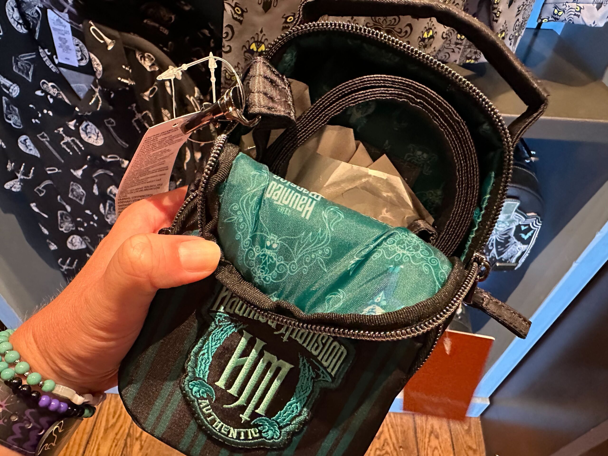 A hand holds a small black and teal handbag with a Haunted Mansion theme, showing an open top with the logo visible inside.