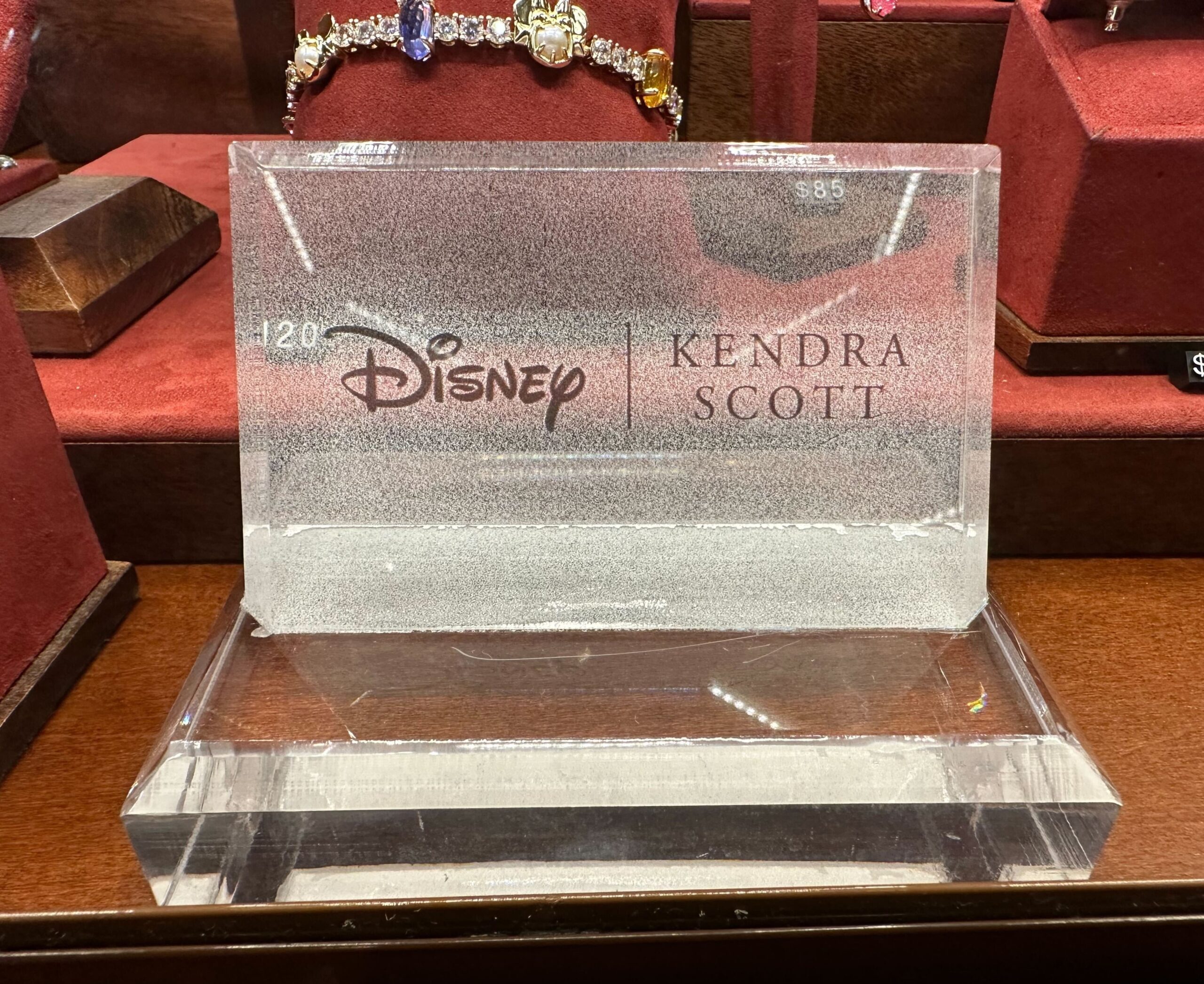 A clear display case contains a Kendra Scott x Disney jewelry piece, priced at $85. The background showcases additional Kendra Scott jewelry items.