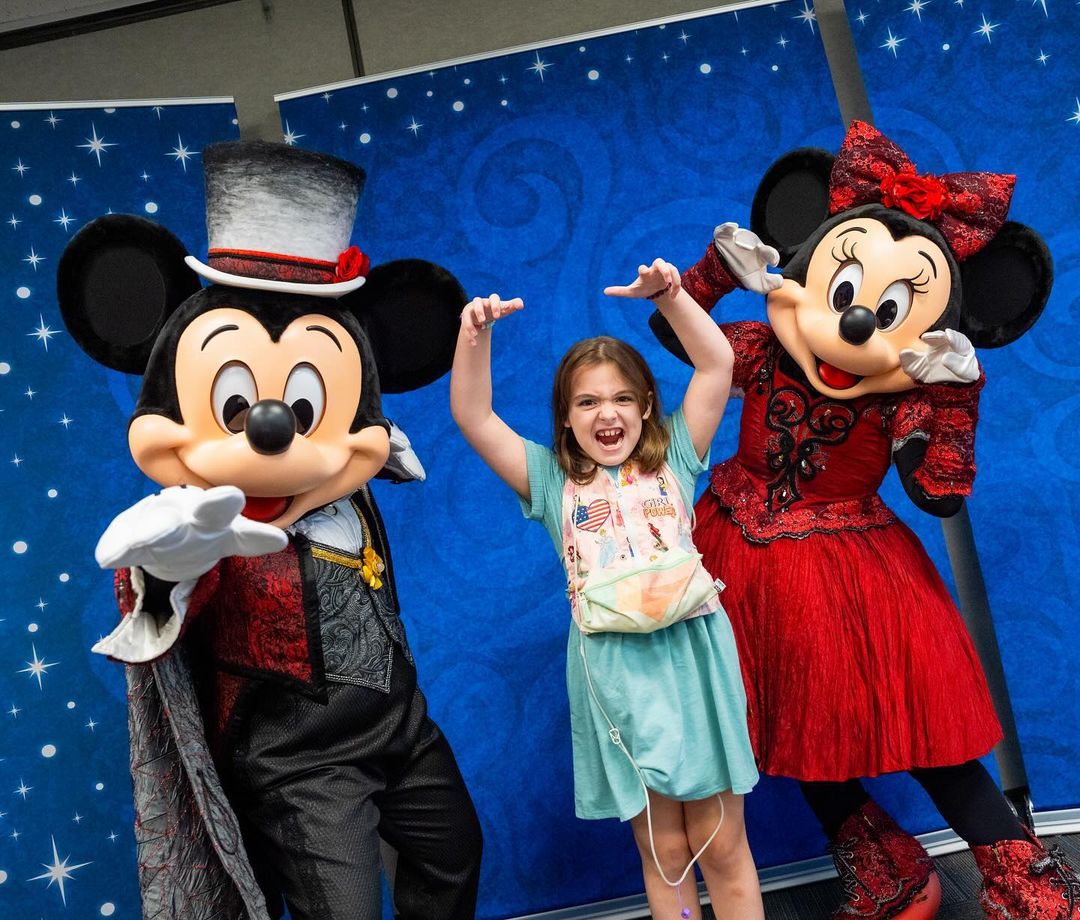 A child poses happily with two people dressed as classic cartoon mouse characters, one in a top hat and the other in a red dress, against a blue backdrop with stars.