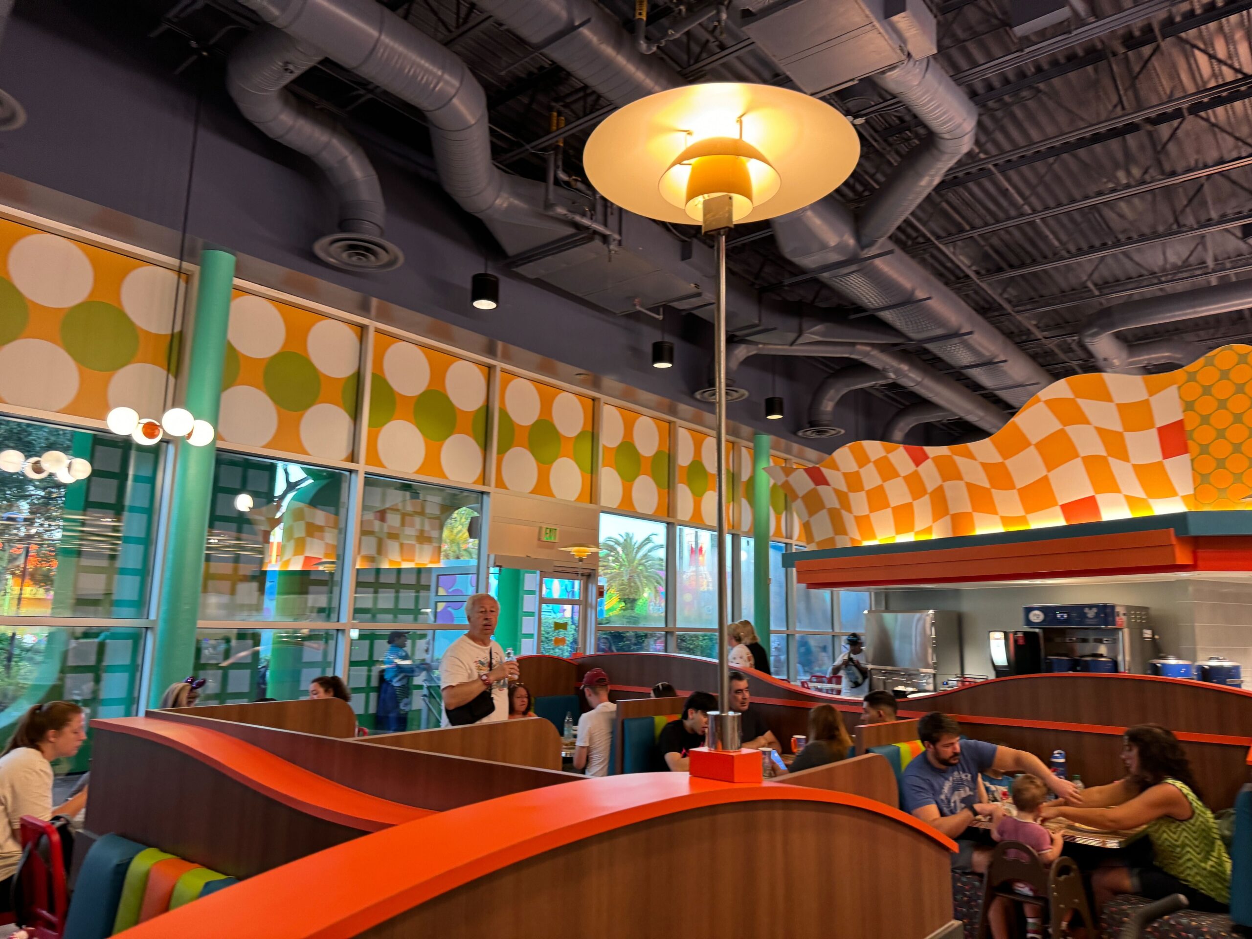 People sitting and standing inside a retro-themed diner with colorful patterned walls, booth seating, overhead lighting, and visible ceiling ducts.