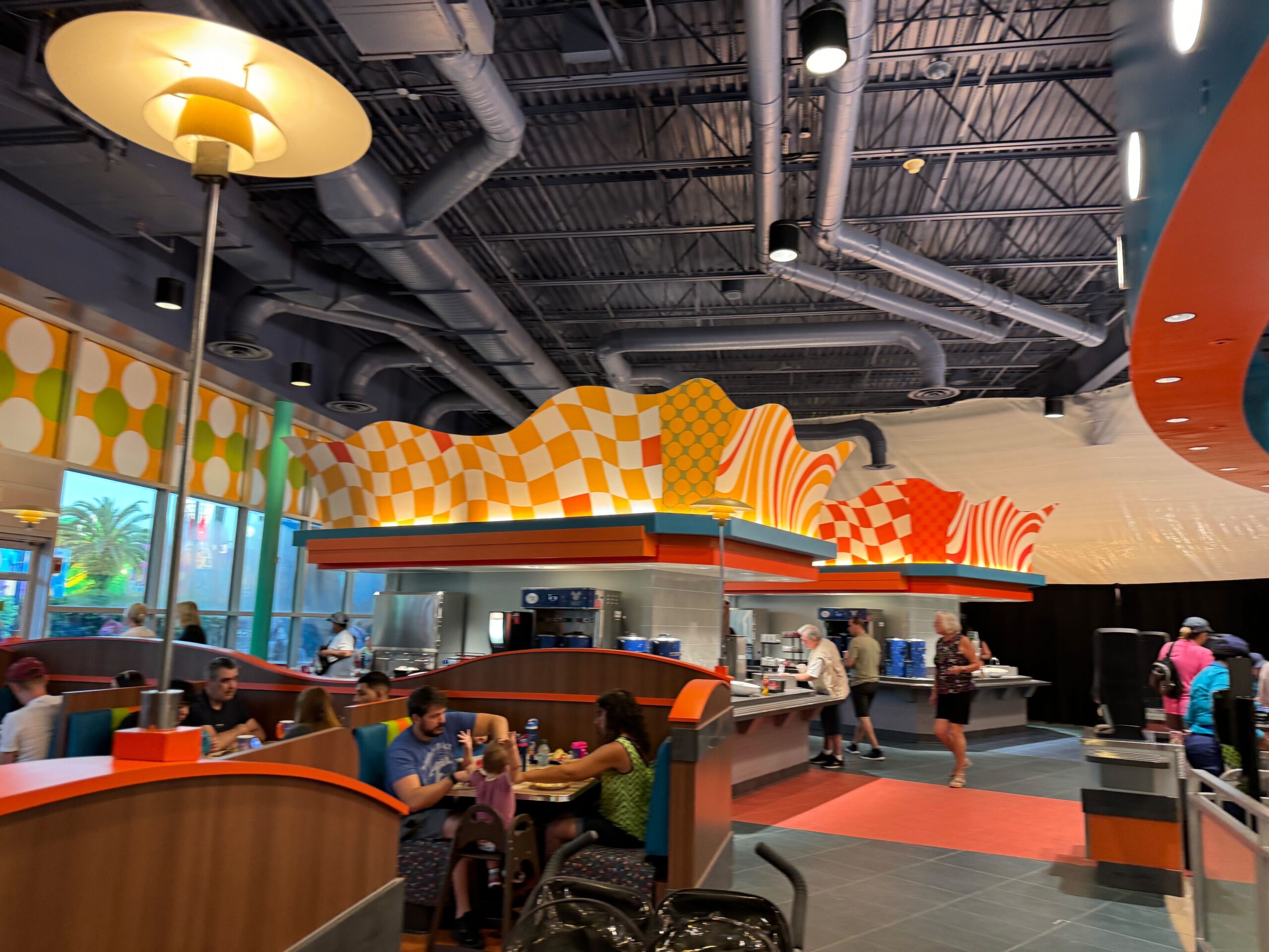 People sitting and eating at colorful, modern-designed restaurant with overhead lights and exposed pipes. Bright, geometric-patterned decorations adorn the space above the food counters.