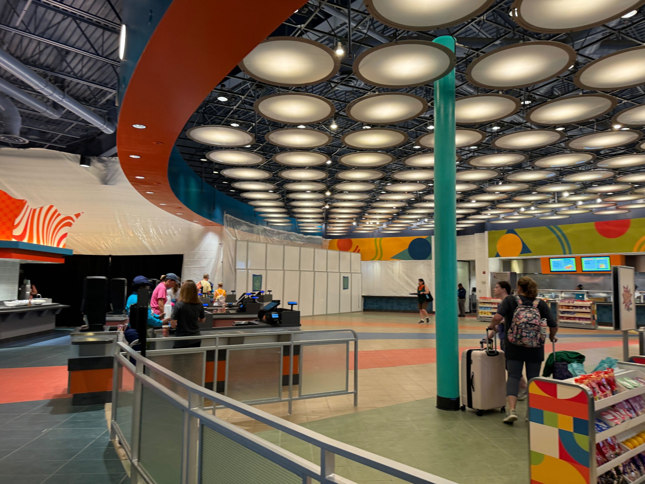 A brightly lit indoor space with a patterned ceiling, various shopping counters, and several people walking or waiting with luggage.