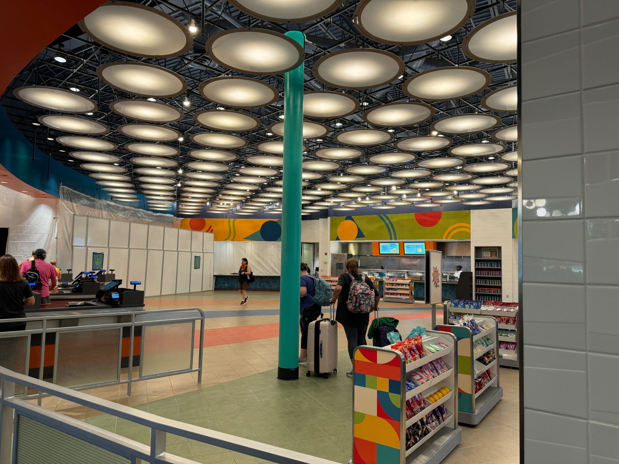 A modern, brightly lit airport or train station terminal featuring a snack shop, informational screens, and passengers with luggage.