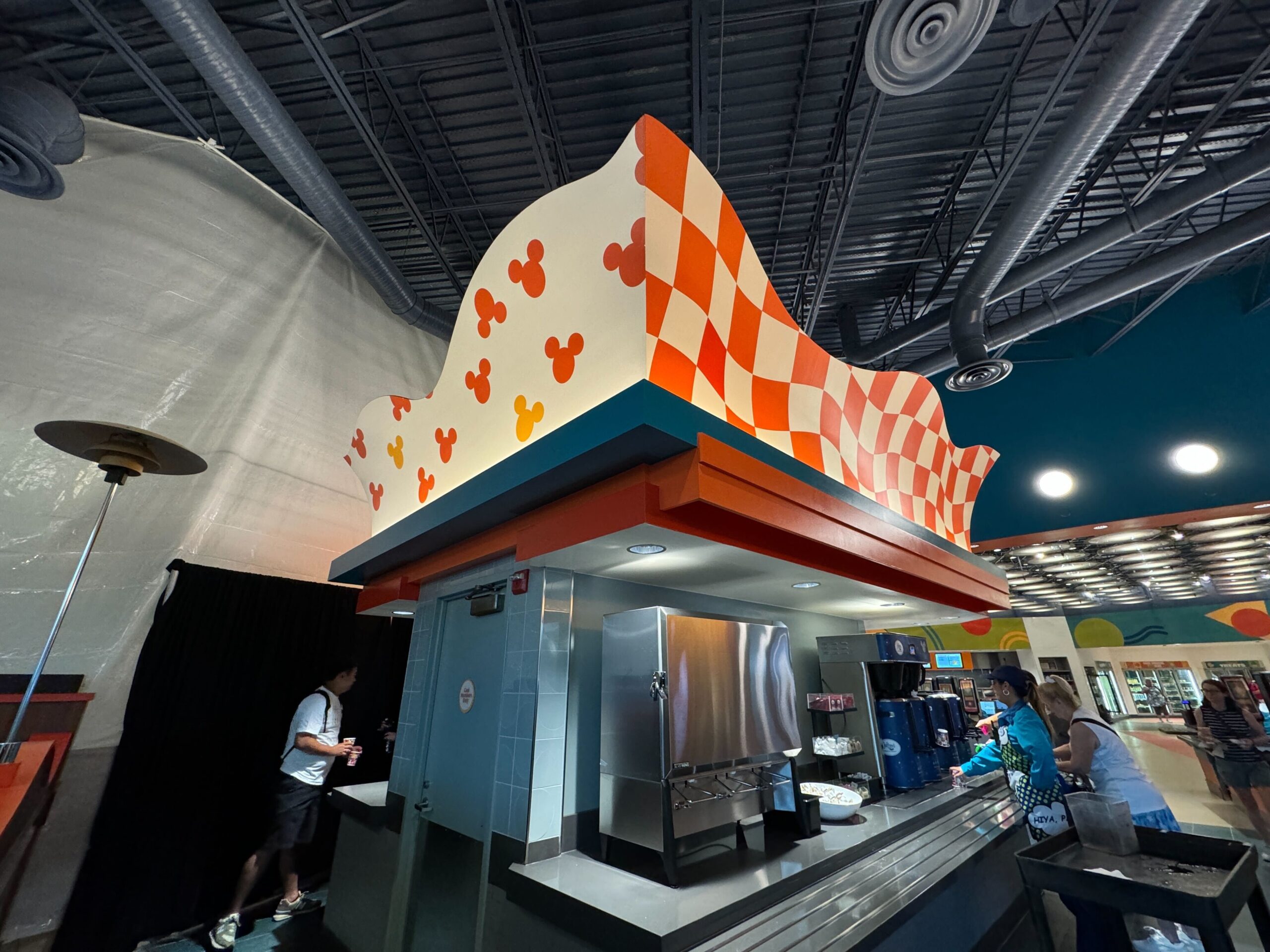 A fast-food counter with an orange and white checkered roof featuring Mickey Mouse silhouettes. People are being served food under bright interior lighting.