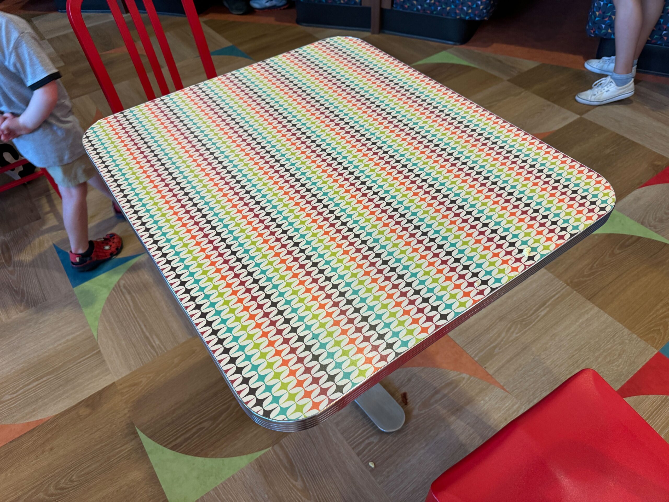 A square table with a colorful, geometric pattern on the tabletop is shown in a dining area with a wooden floor. Nearby, a child's legs and feet are visible.