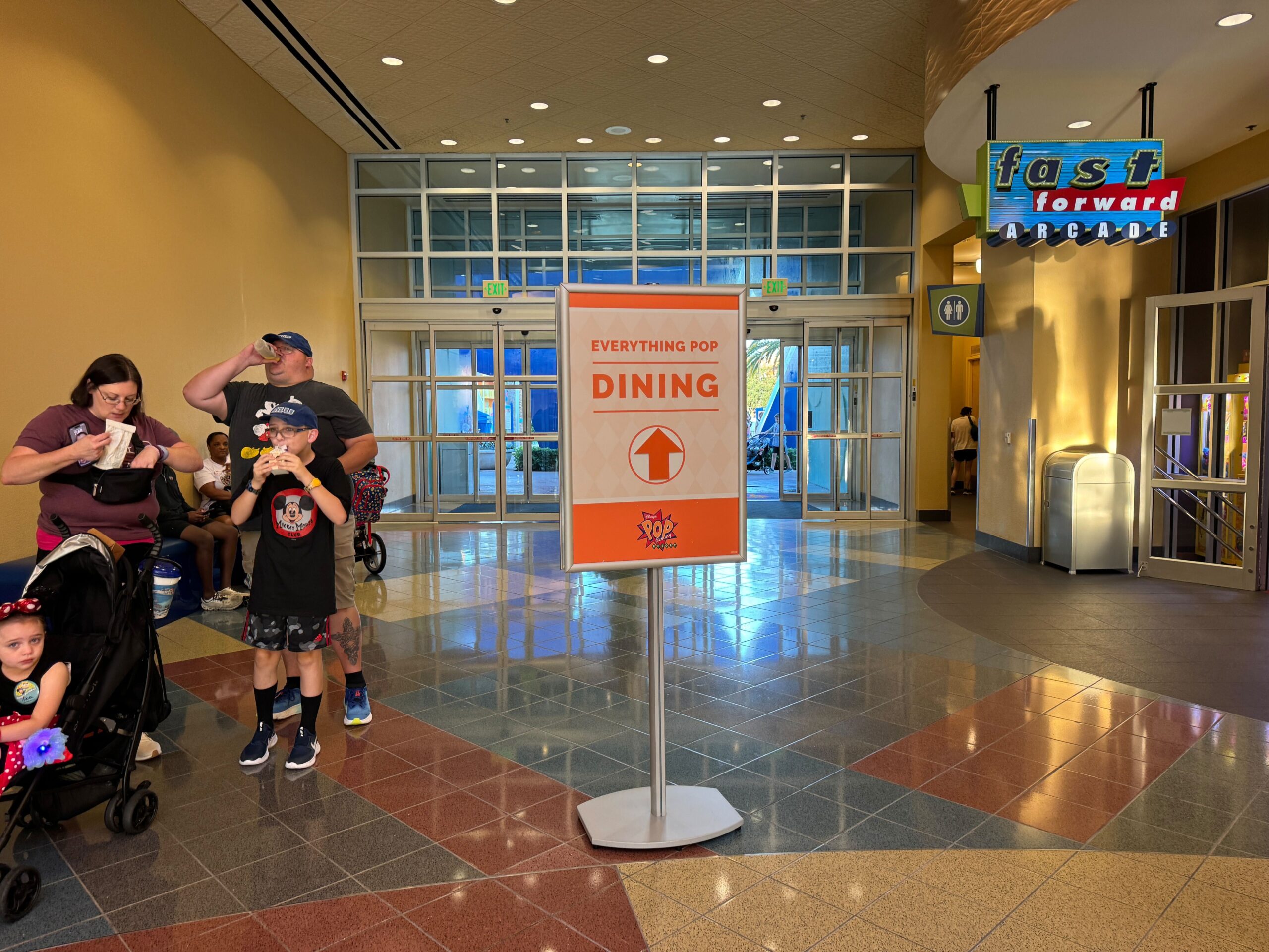 A sign in a lit hallway directs to "Everything Pop Dining" with families and children nearby. An arcade named "Fast Forward" is on the right.