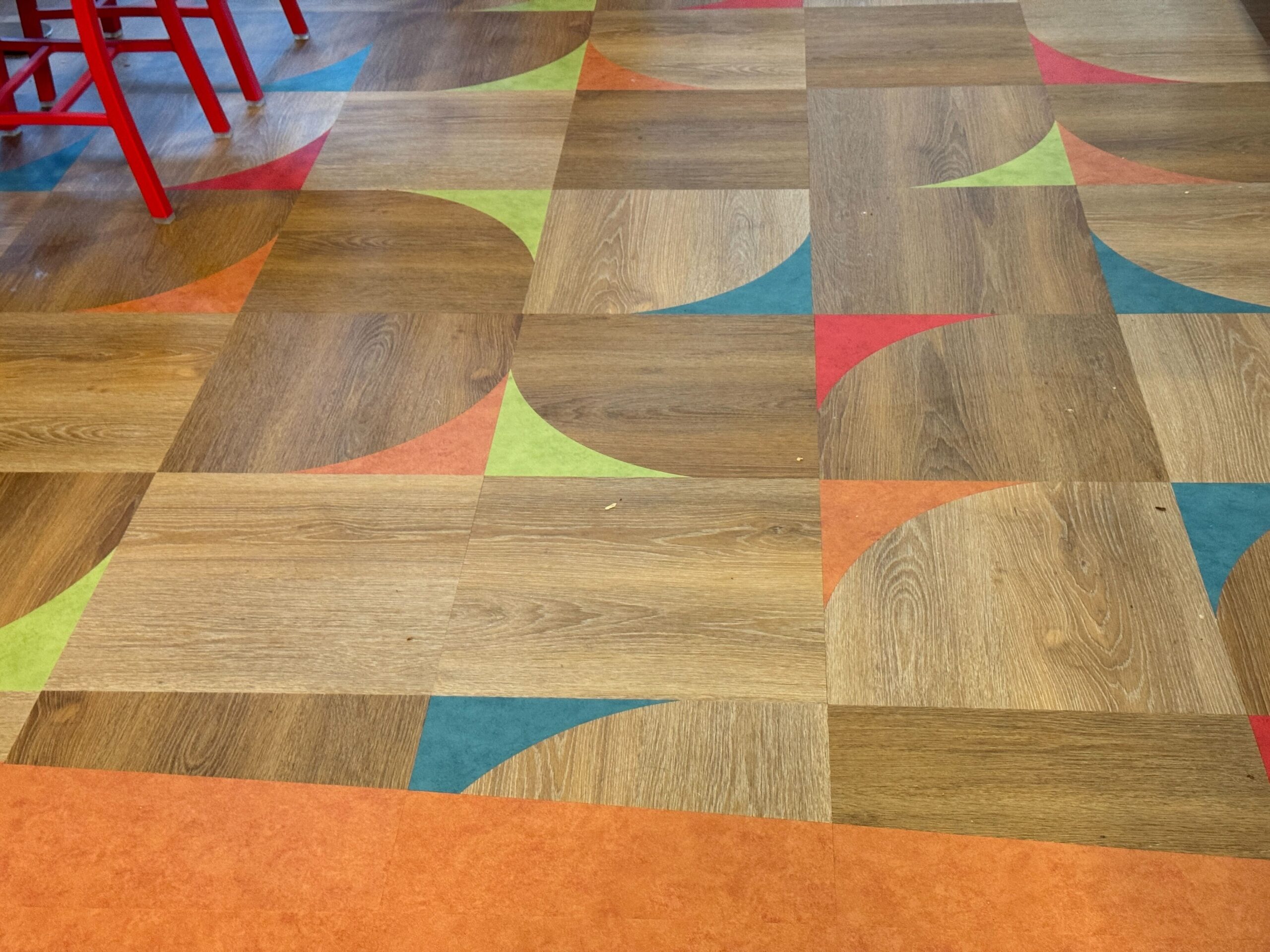 A patterned wooden floor with multicolored geometric designs in orange, red, green, and blue sections. Red chair legs are partially visible at the top left corner.
