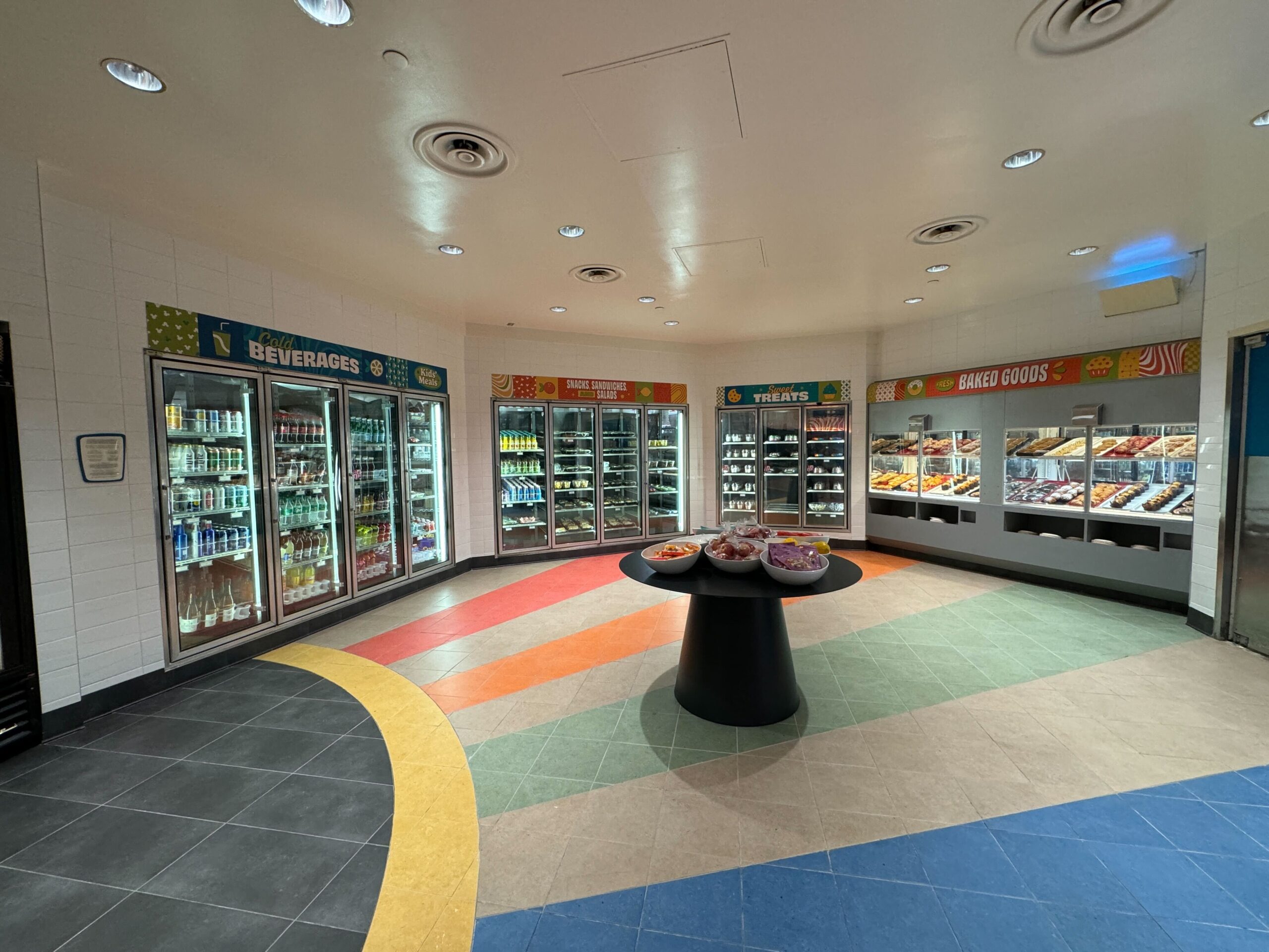 A brightly lit convenience store section with stocked refrigerated shelves for beverages and snacks, and a central table displaying various fruit bowls.