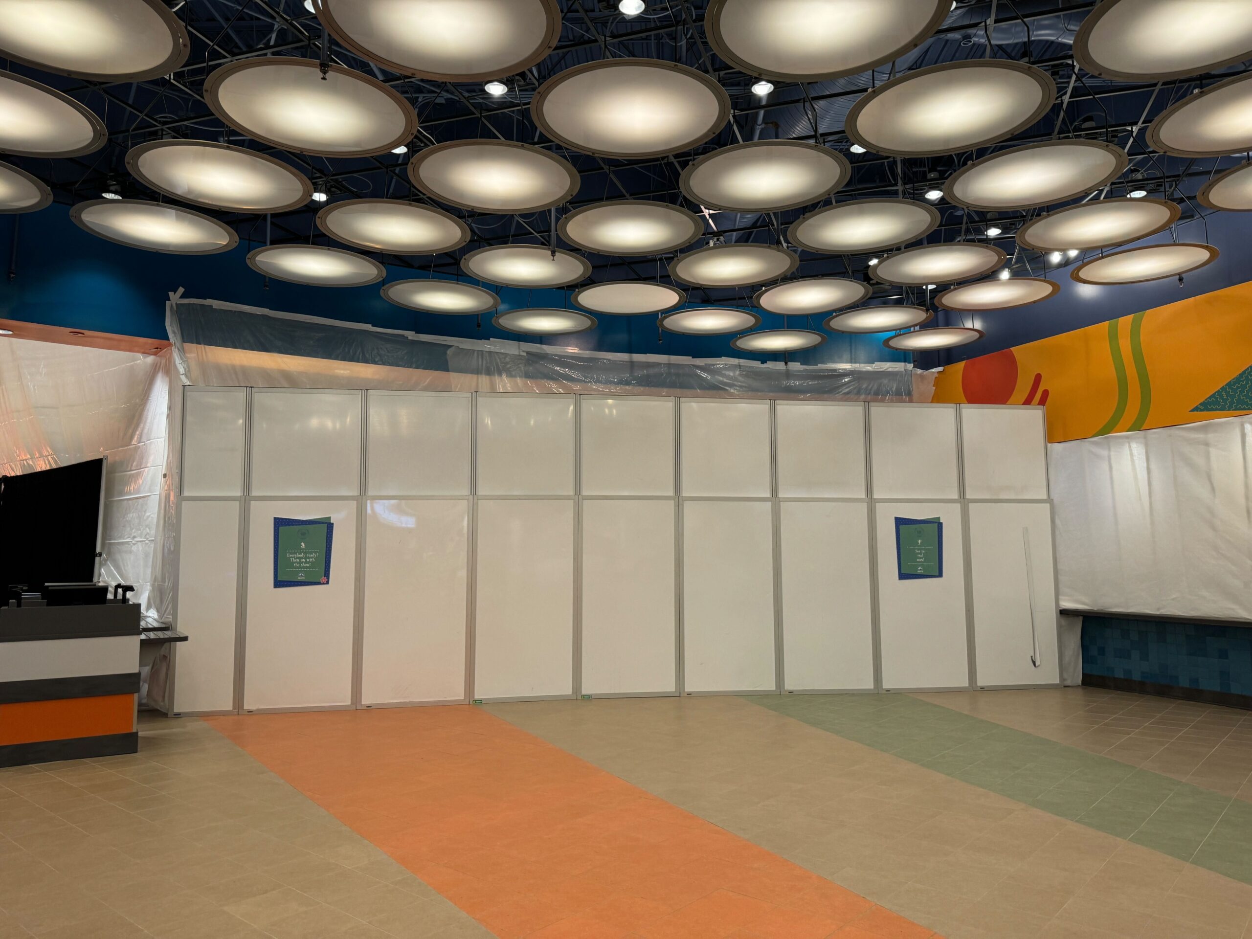 A white temporary construction wall with two signs is set up in an indoor area with round ceiling lights and multicolored flooring.
