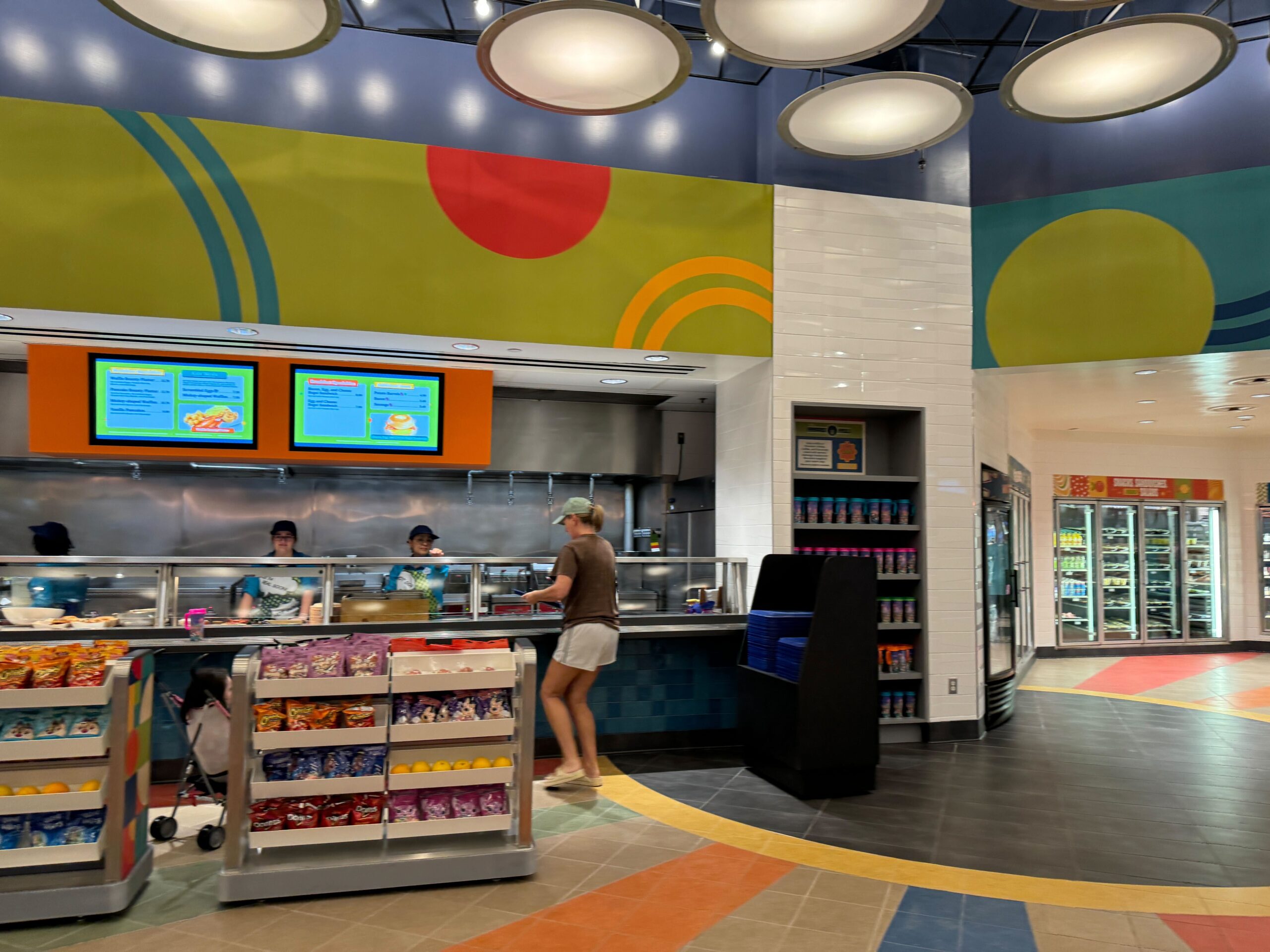 A person stands near a counter with food items and snacks in a brightly colored cafeteria with staff behind a serving area under menu screens.