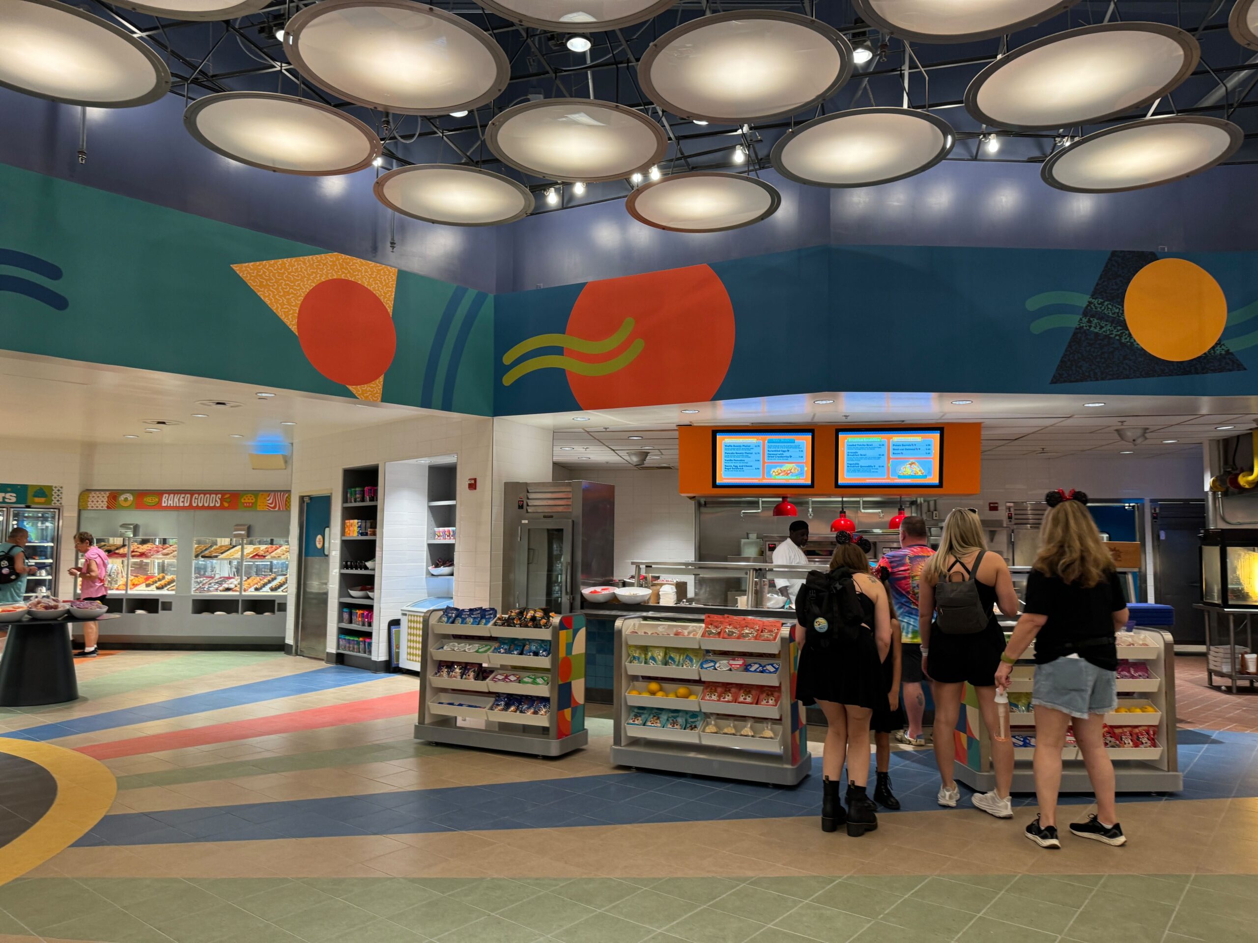 People standing in line at a colorful, modern cafeteria with illuminated circular ceiling lights and various snacks displayed on shelves.