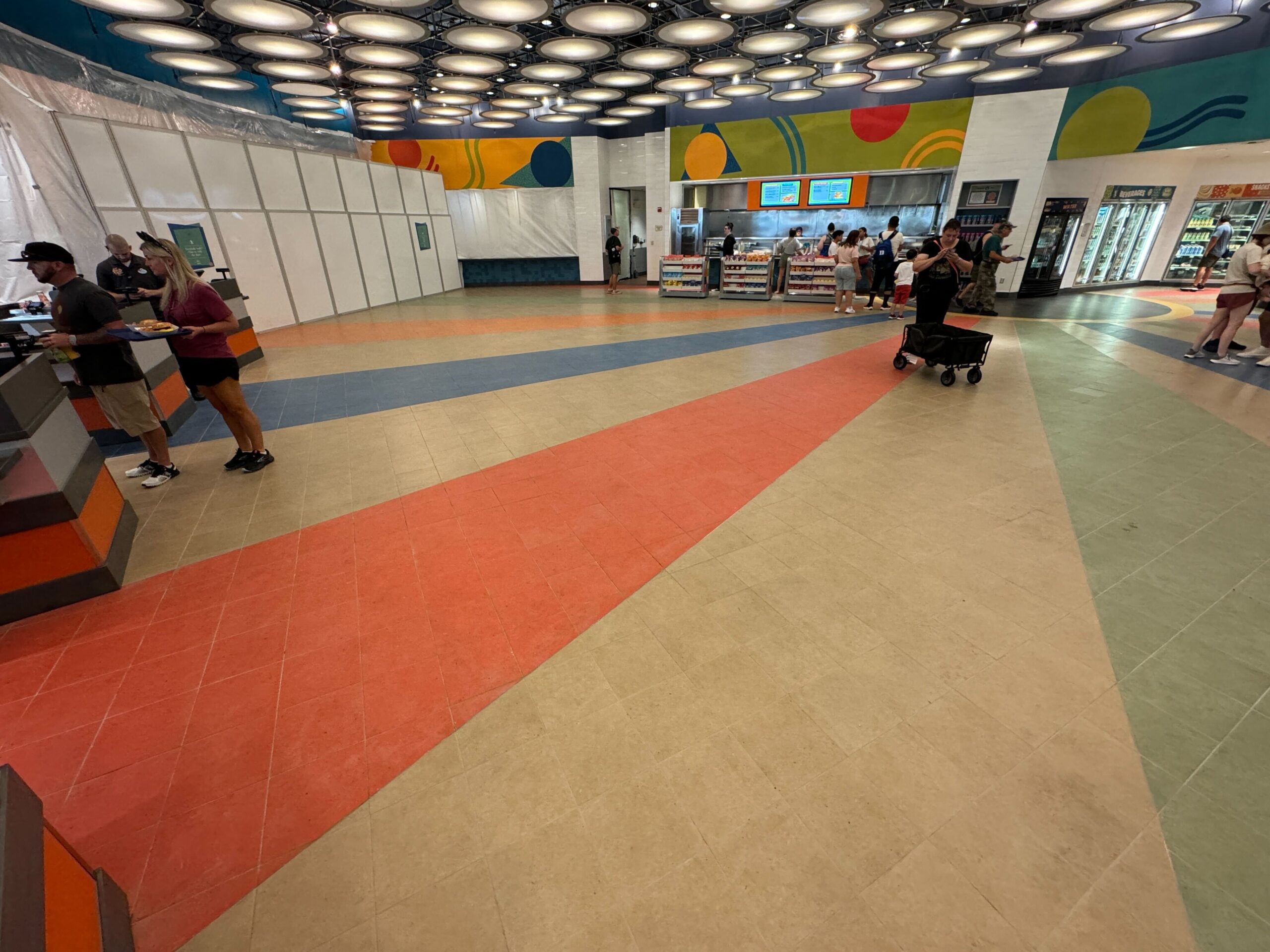 A bright, colorful indoor food court featuring geometric floor patterns, several food service counters with patrons, and a refrigerated drinks section on the right. This lively scene is reminiscent of the vibrant atmosphere at Everything Pop Food Court.