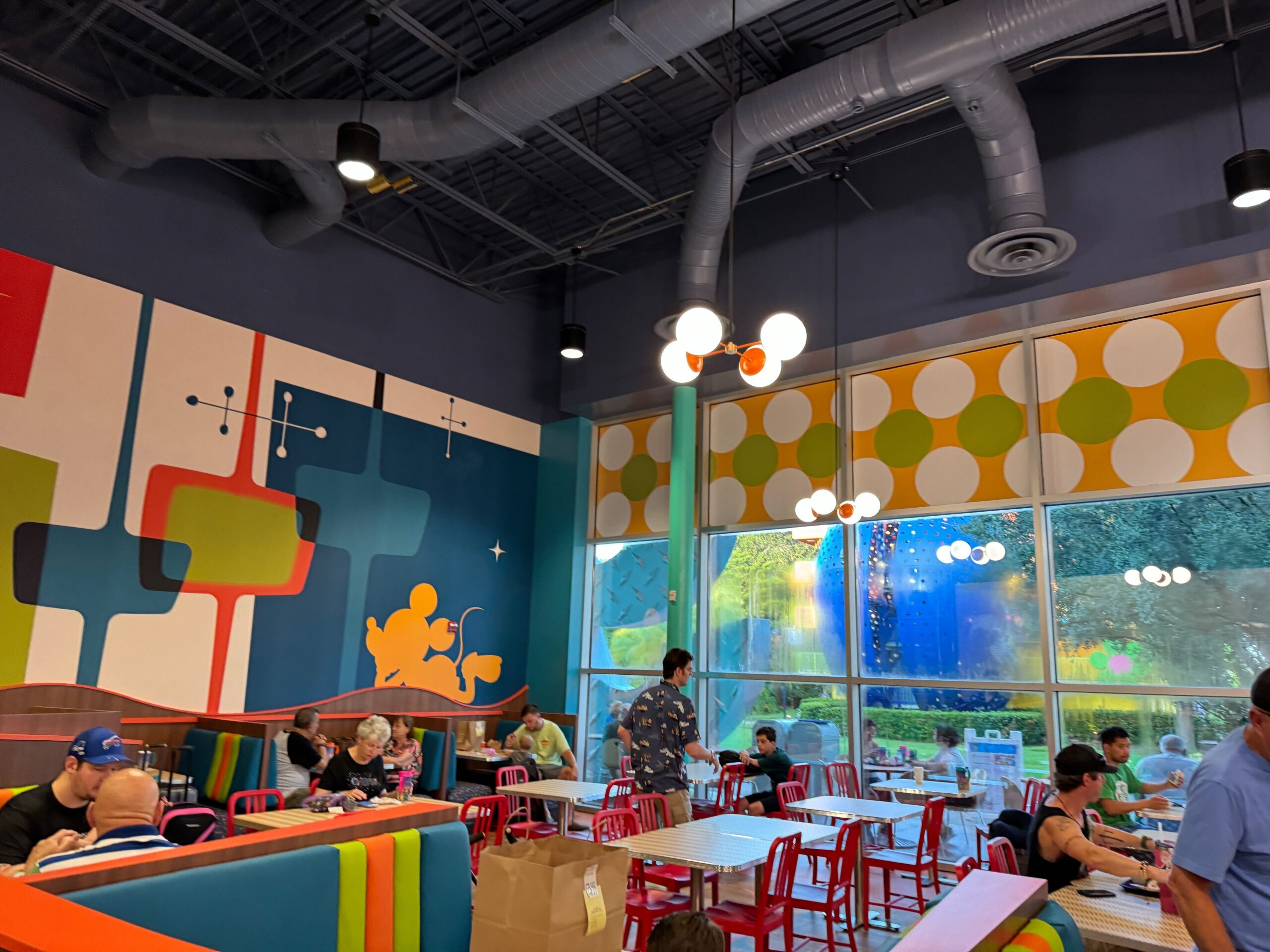 Colorful restaurant interior with modern decor, featuring geometric patterns on the walls and windows. Patrons sit at tables, a few standing and moving around. Overhead lighting and exposed ceilings are visible.