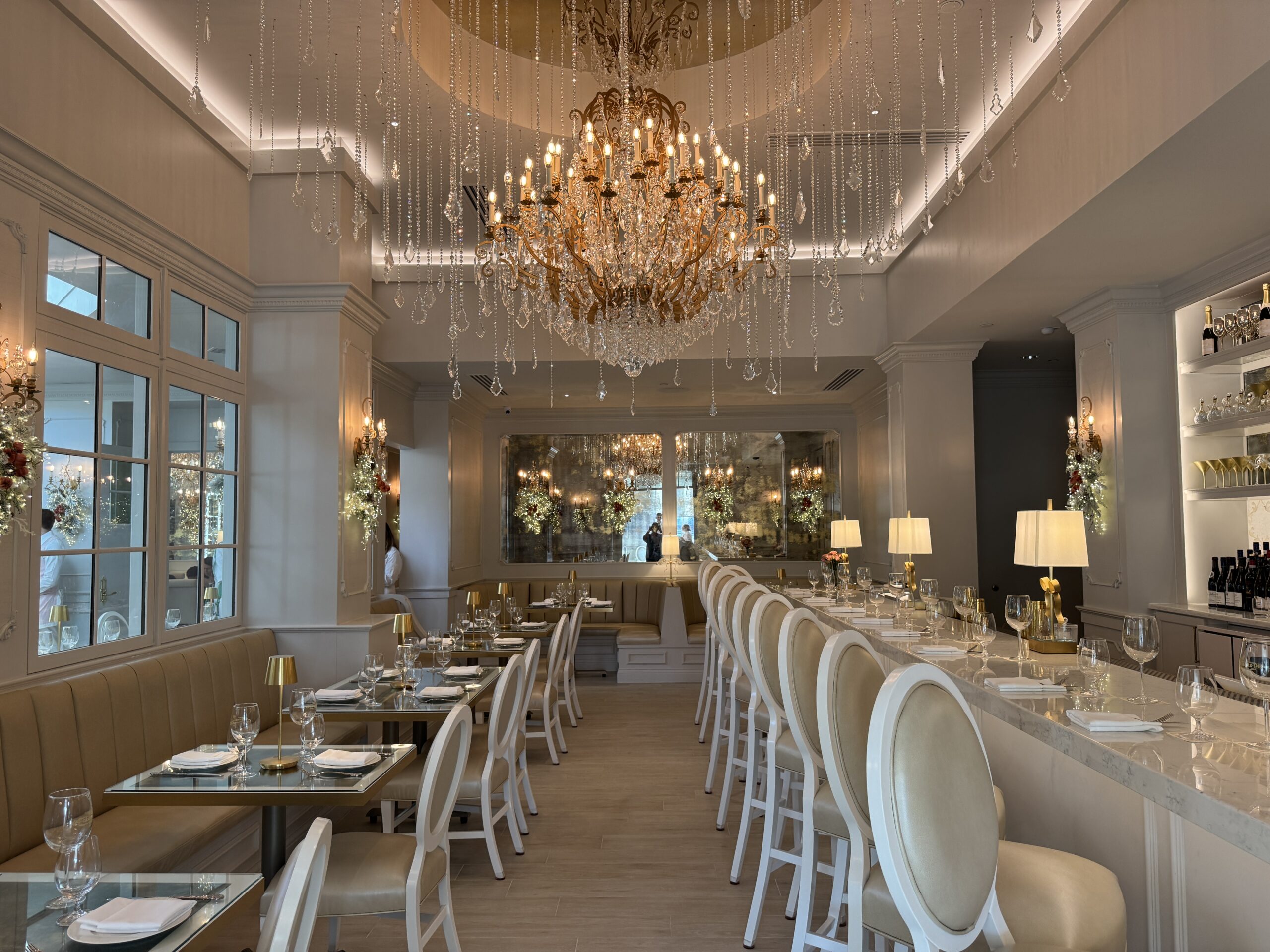 The elegant restaurant interior, reminiscent of a sophisticated cake bake shop, features a large chandelier, white chairs, and set tables. Mirrors reflect the warm, inviting atmosphere where reservations are recommended for an unforgettable dining experience.