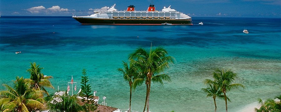A Disney Cruise Line ship glides on a clear blue ocean near a tropical island with palm trees and a sandy beach.