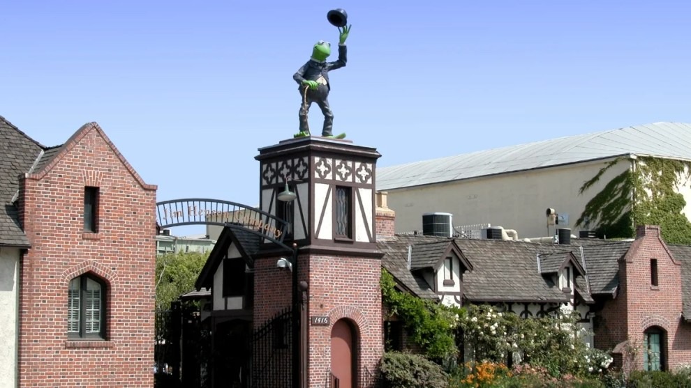 A statue of a frog in a suit and hat, reminiscent of Jim Henson Company's iconic characters, stands atop a brick building with a clear blue sky in the background.