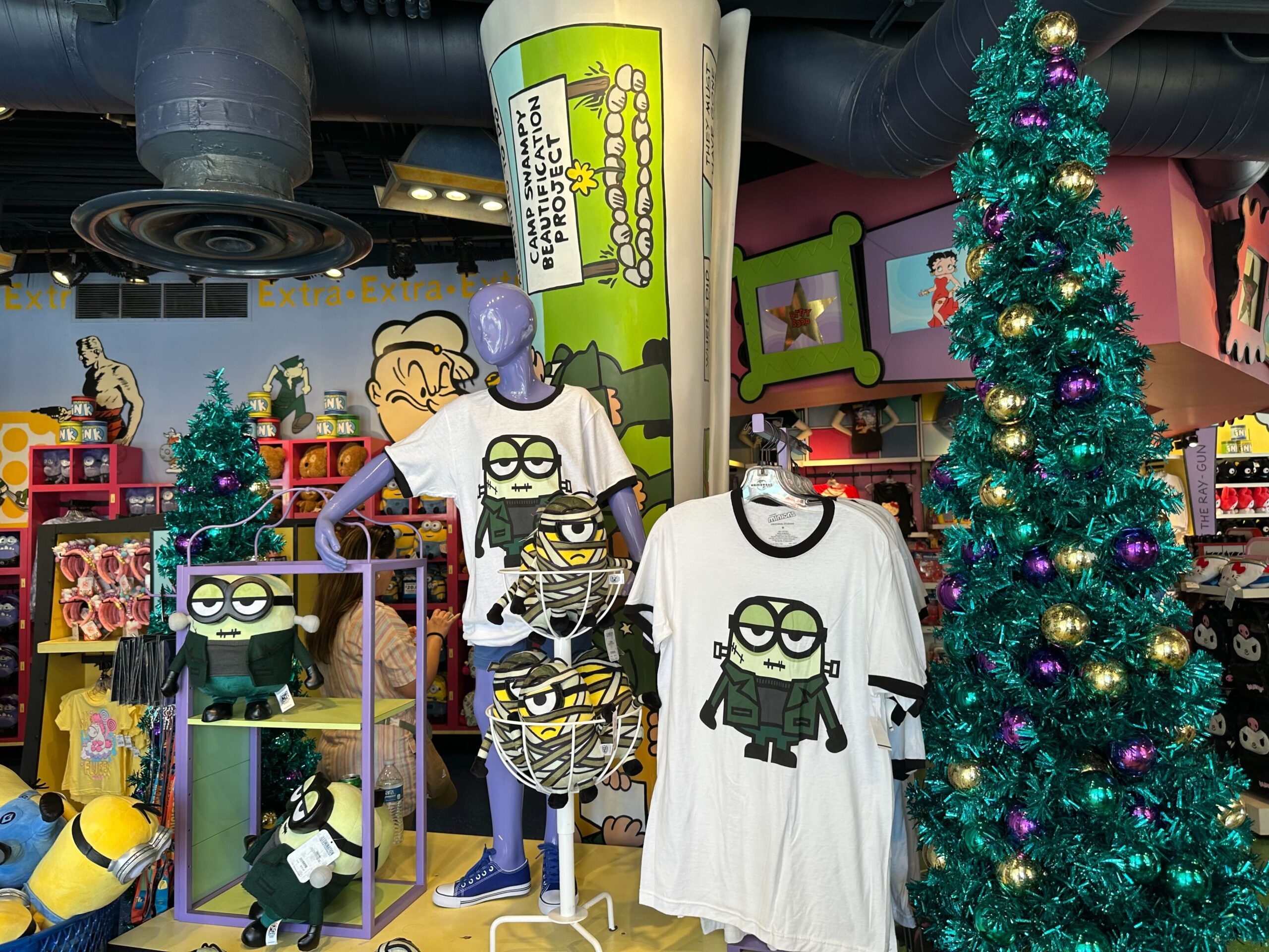 A store display featuring Minions merchandise includes t-shirts, plush toys, and green tinsel Christmas trees.