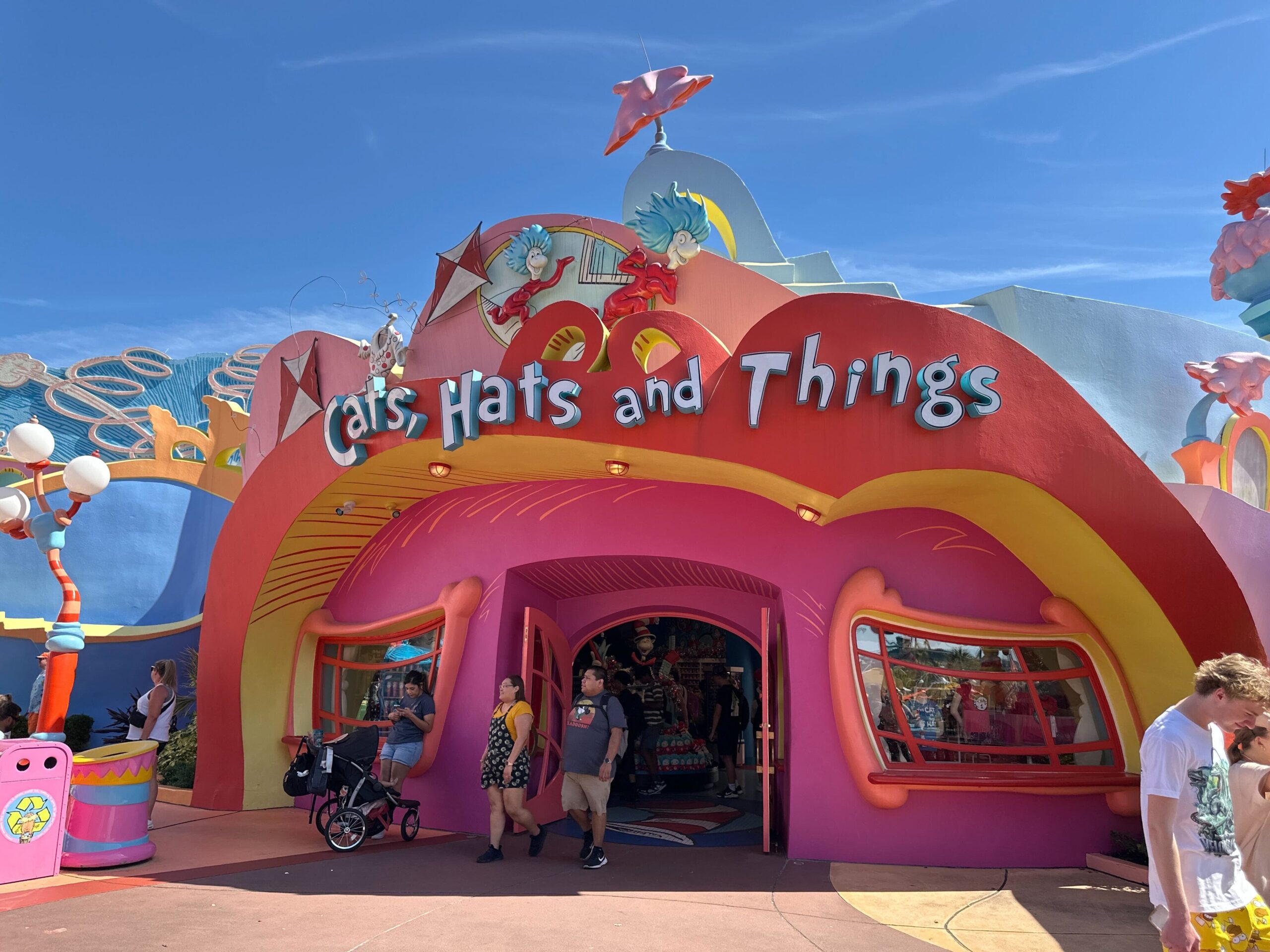People walk past a colorful "Cats, Hats and Things" store with playful designs under a clear blue sky.