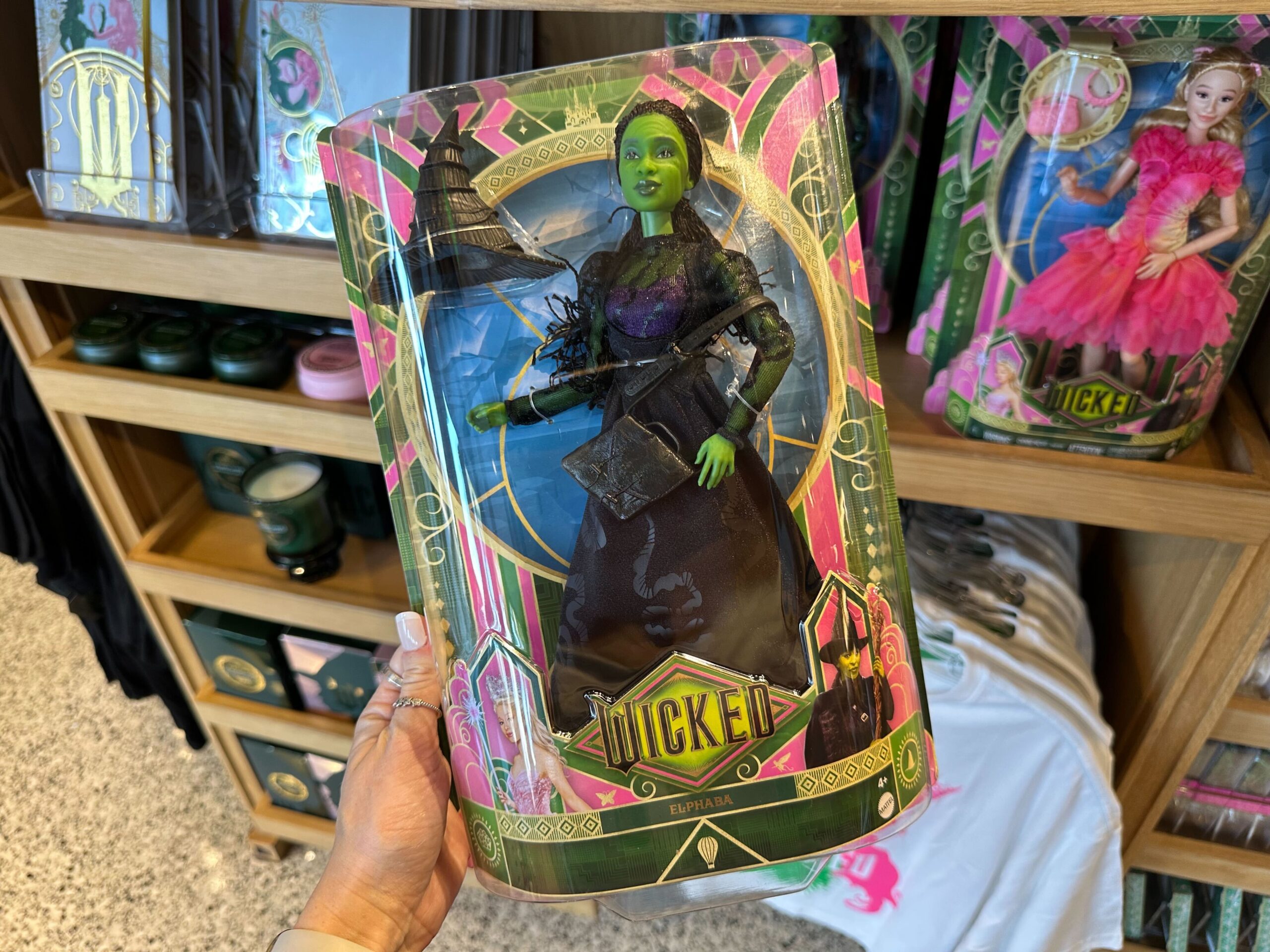 A hand holds a "Wicked" themed doll in green and black attire, packaged in a decorative box. This exclusive Wicked Merchandise is perfect for any fan visiting Universal Orlando Resort. Shelves with similar enchanting items are visible in the background.