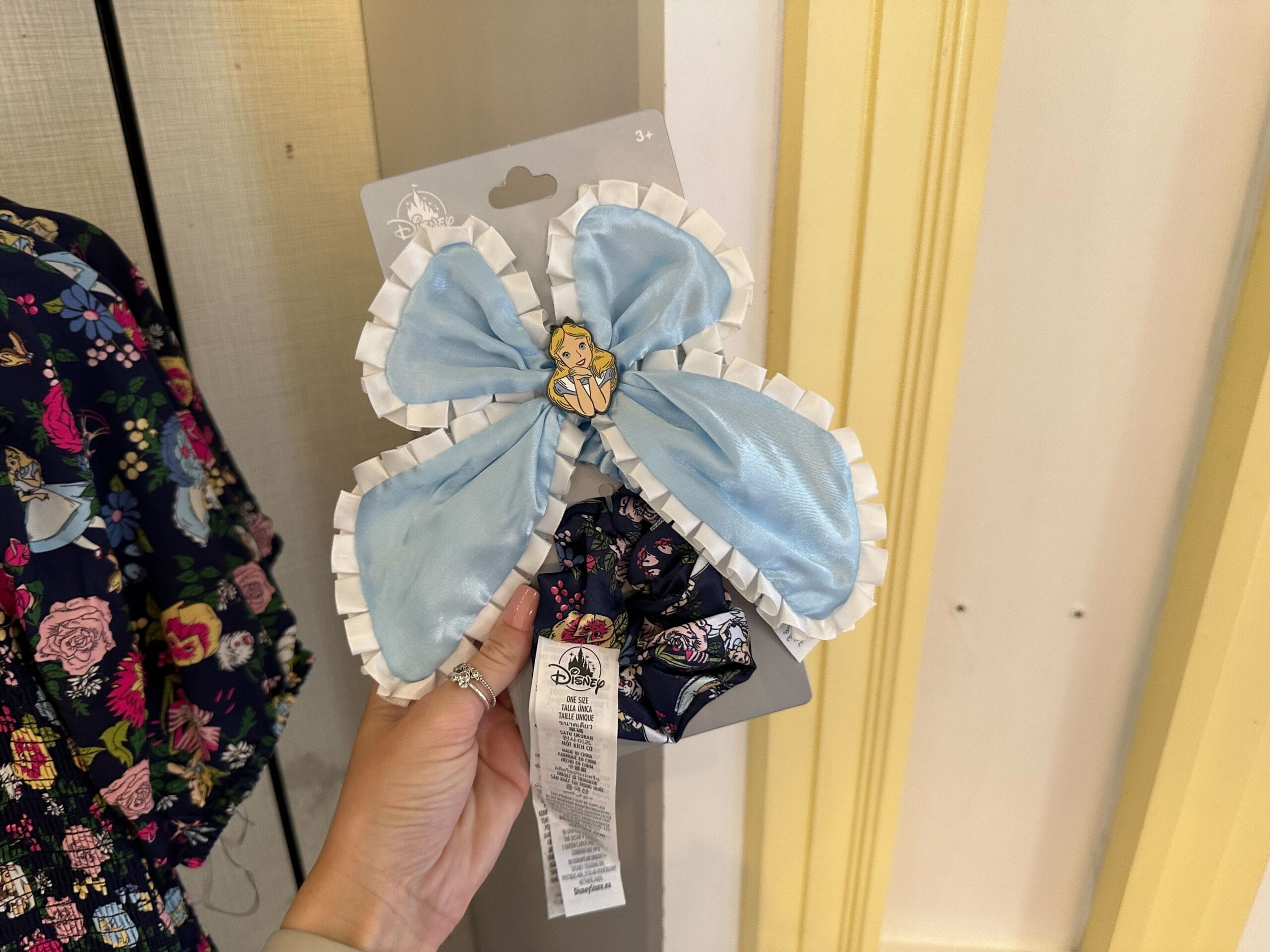 A hand holds a package with a blue hair bow adorned by an Alice in Wonderland cartoon character. The background showcases fabric with a floral pattern, adding whimsical charm.