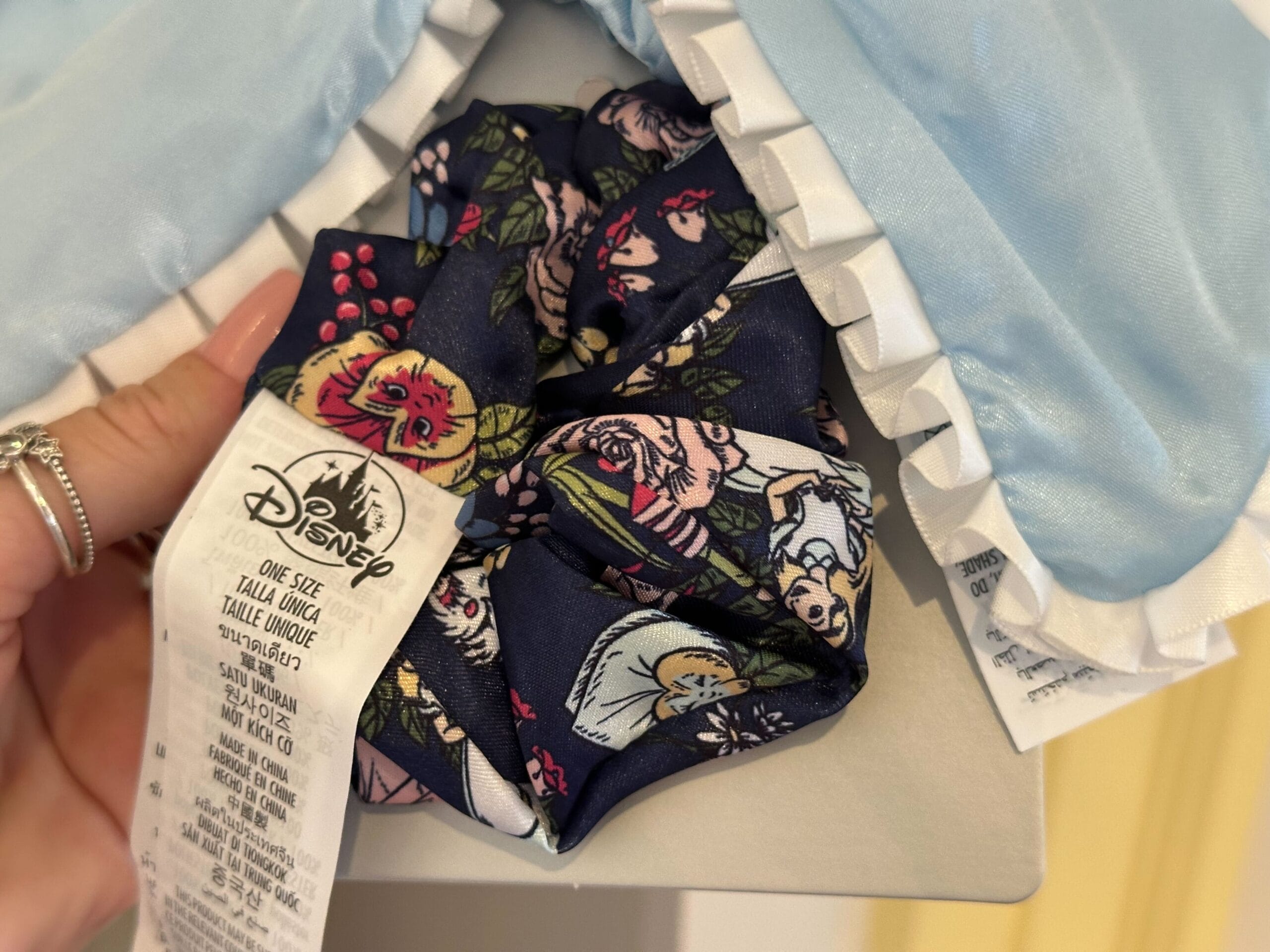 Close-up of a Disney-themed scrunchie featuring Alice in Wonderland floral and character designs, held by a hand adorned with rings. A white tag displays brand and size information.