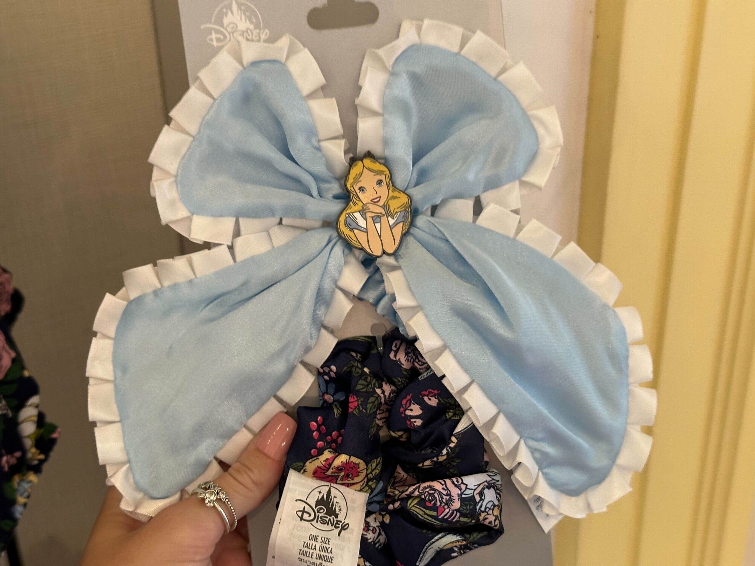 A light blue bow with a ruffled white edge showcases a pin of an animated character reminiscent of Alice in Wonderland at the center. It is elegantly displayed alongside a dark patterned fabric.