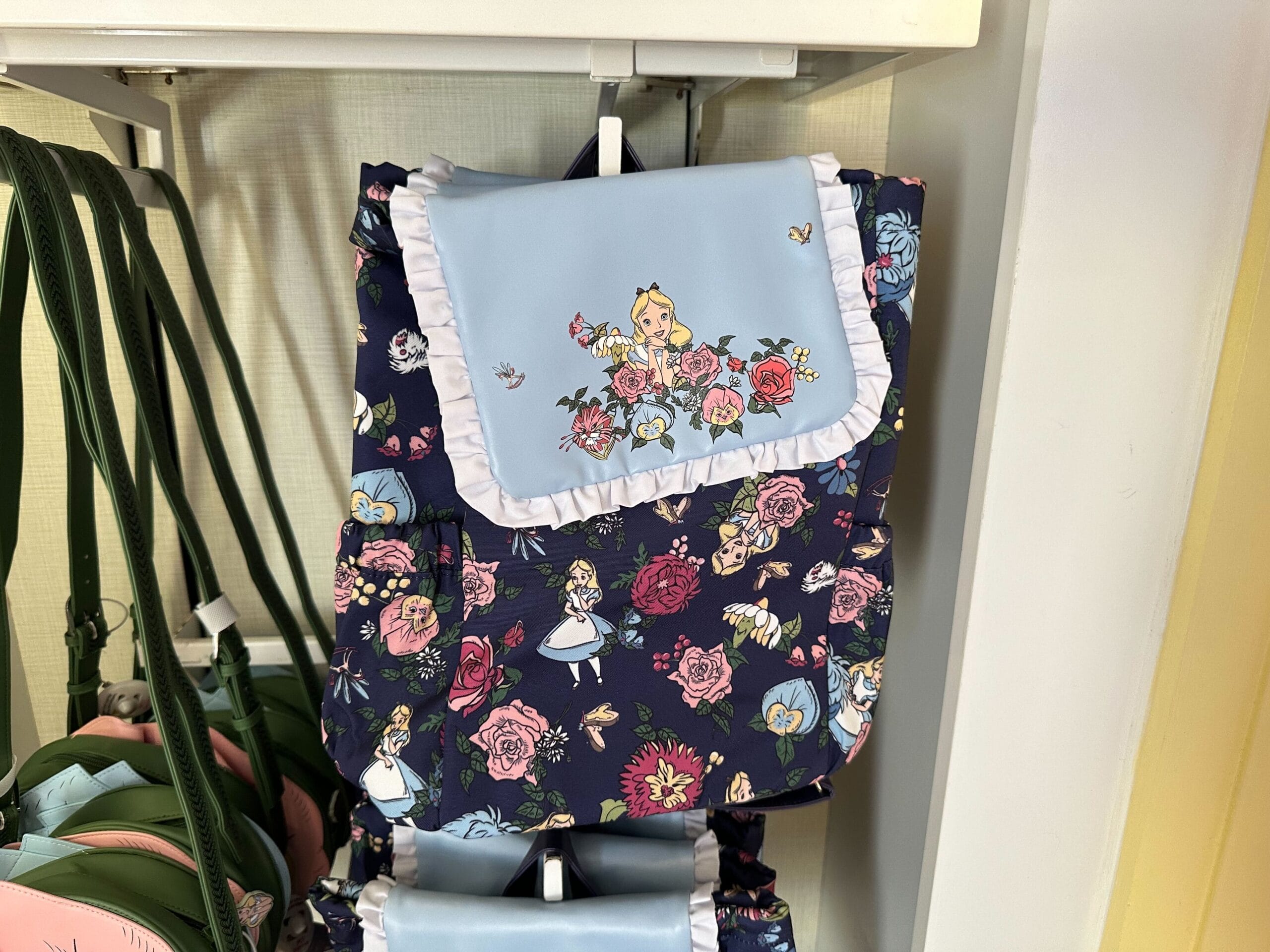 A colorful backpack featuring floral designs and an Alice in Wonderland character illustration on the flap, displayed on a store shelf, inspired by Ashley Eckstein's whimsical style.
