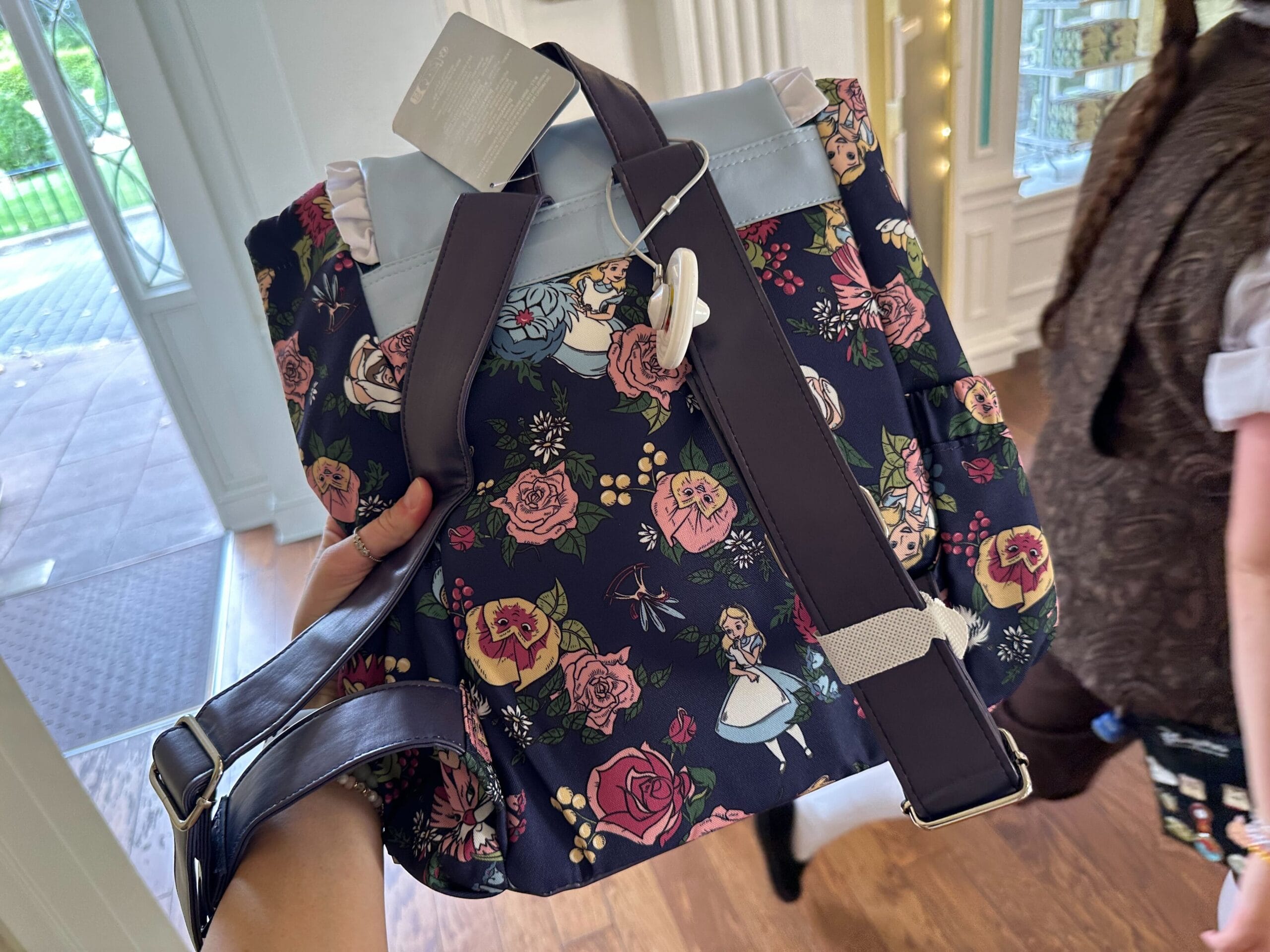 A floral-patterned bag with dark straps is held by a person. The bag, reminiscent of Alice in Wonderland, features illustrations of a girl with an umbrella among the flowers. Another person is walking in the background, creating a whimsical scene.