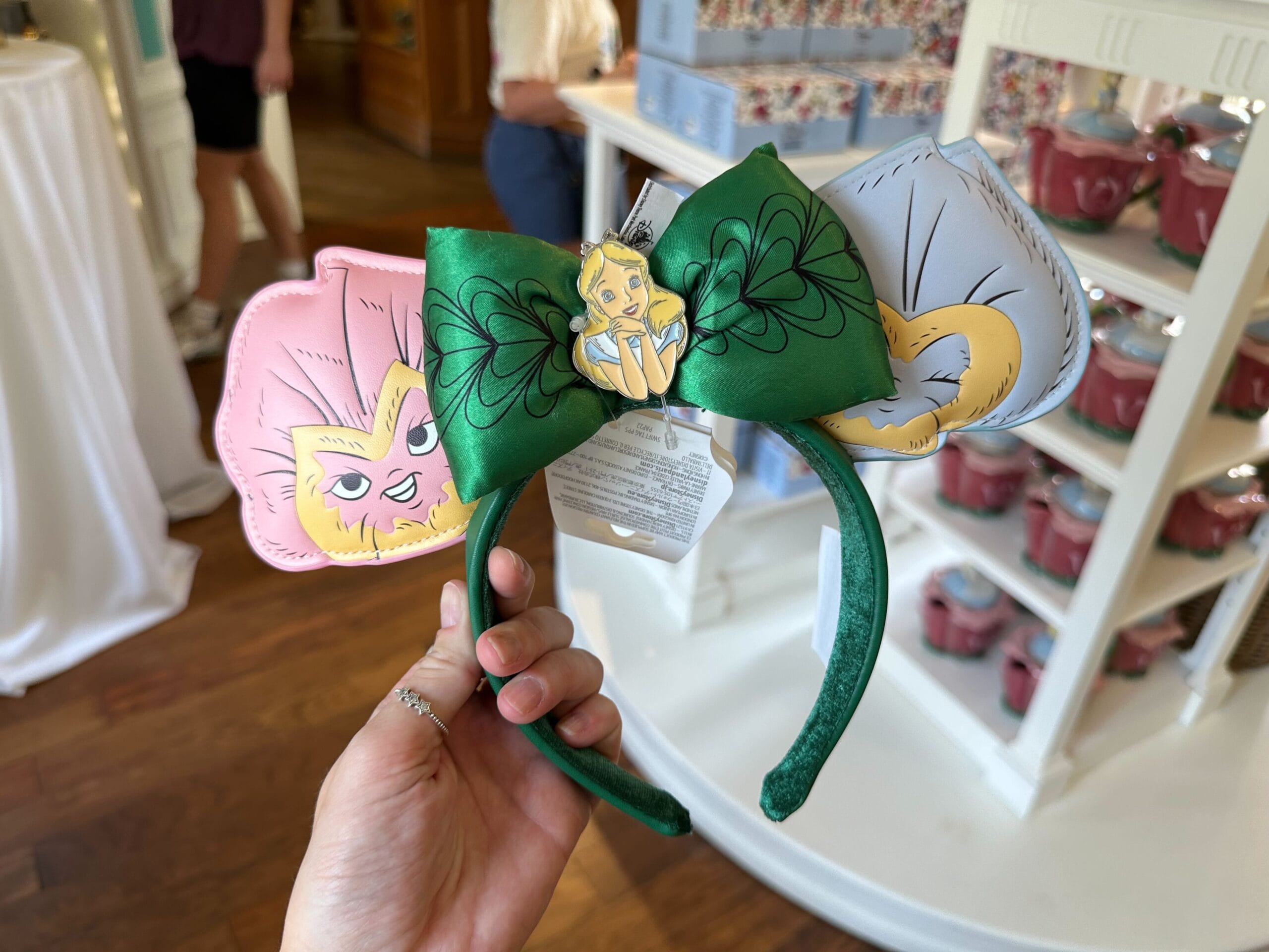 The green headband, adorned with a large bow featuring leaf patterns and a golden character, is reminiscent of Alice in Wonderland. It’s flanked by charming animal faces on pink and gray ears. In the background, shelves brim with various whimsical items.