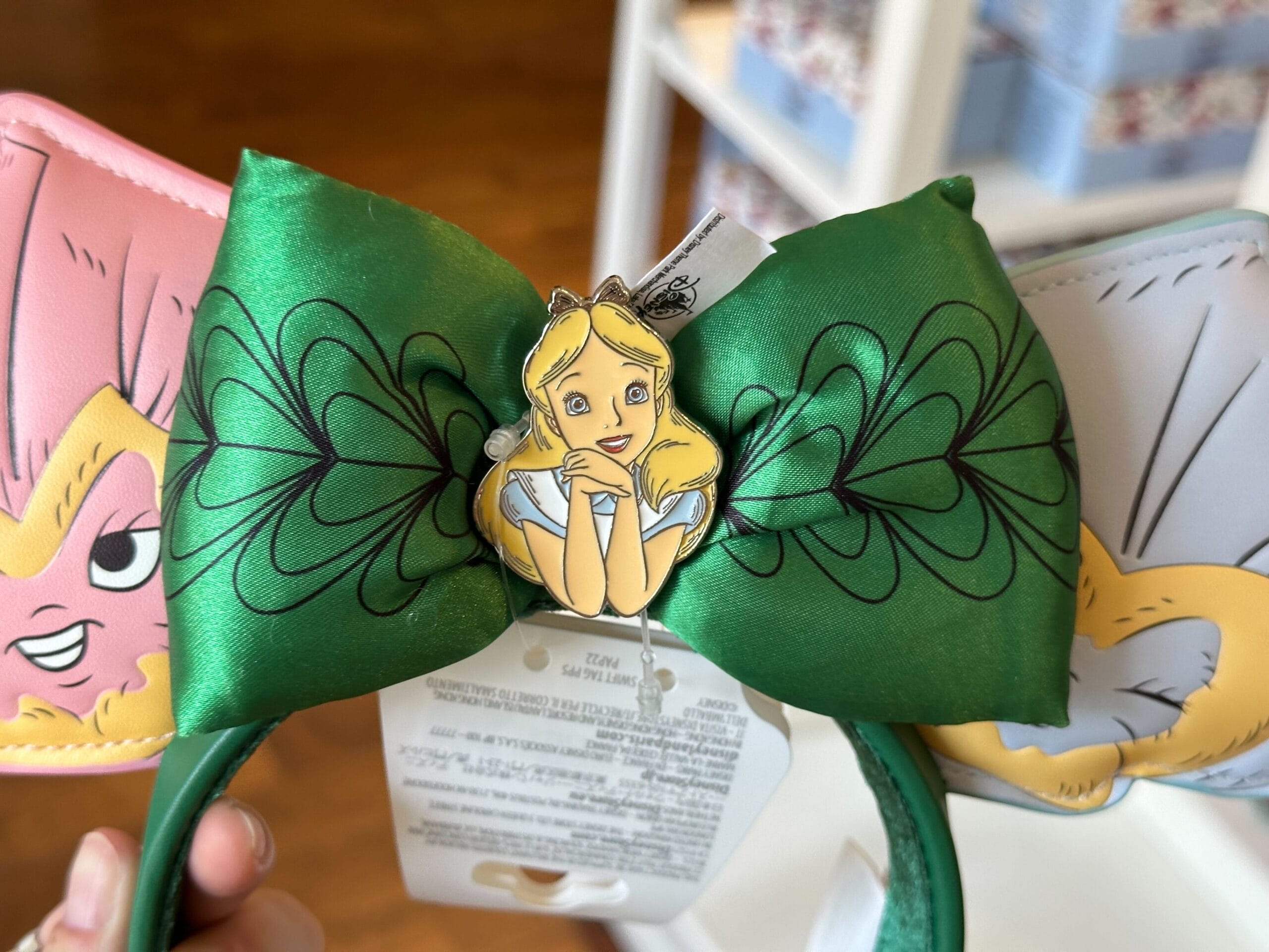 Green headband with a large bow featuring a cartoon character reminiscent of Alice in Wonderland, sporting blonde hair. The whimsical character is surrounded by leafy designs. This enchanting accessory, available in store, might even catch Ashley Eckstein's eye.