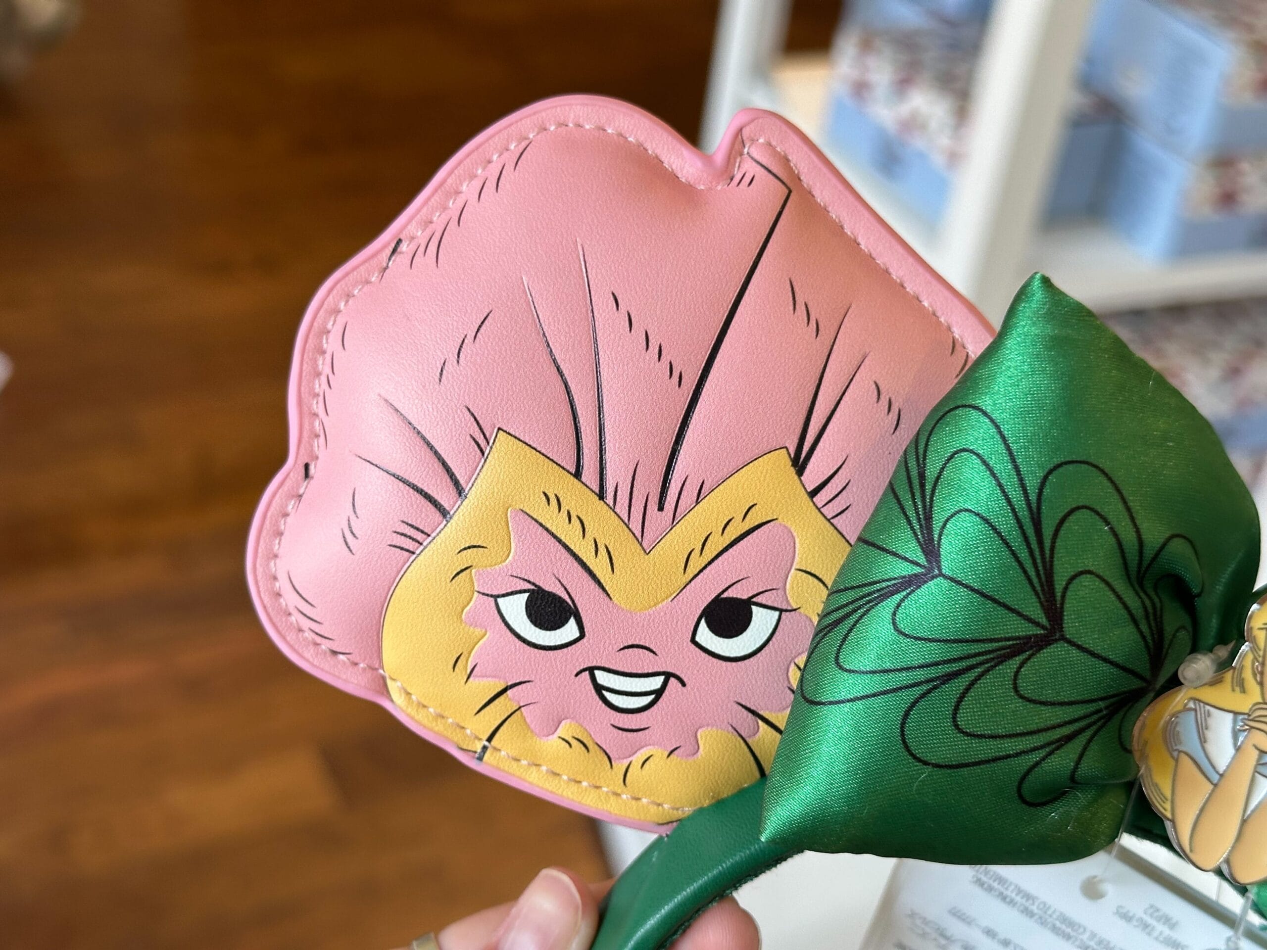 A close-up of a hand holding a flower-shaped pink cartoon character accessory, reminiscent of something from Alice in Wonderland, with a smiling face next to a green leaf-shaped object in a store setting.