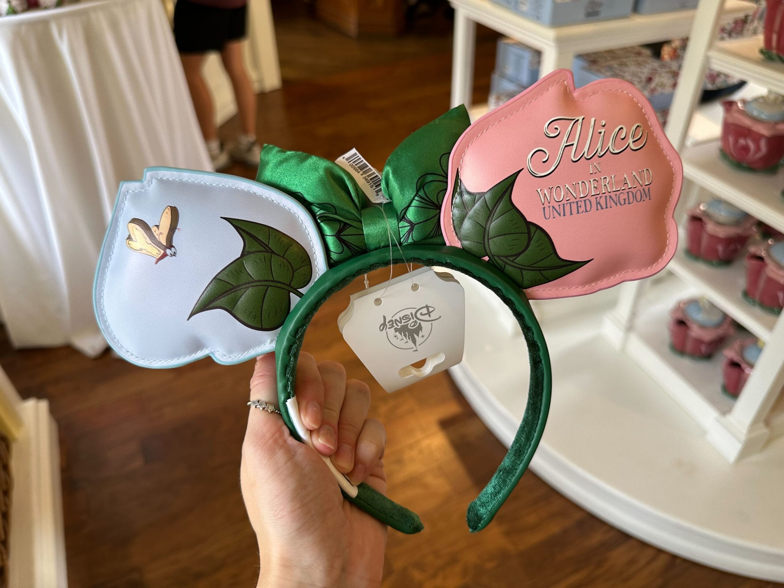 A hand holds a headband adorned with green leaves and flower-shaped decorations inscribed with "Alice in Wonderland United Kingdom," all displayed in a store setting inspired by the whimsical style of Ashley Eckstein.