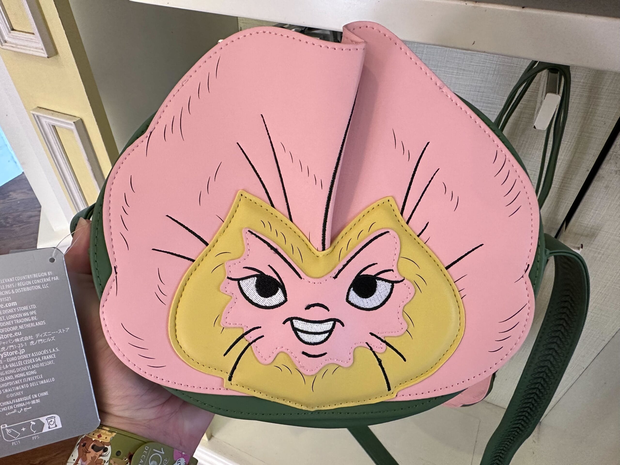 A hand holding a whimsical backpack with a cartoon face design, reminiscent of Alice in Wonderland, featuring vibrant pink and yellow colors, resting playfully on a shelf.