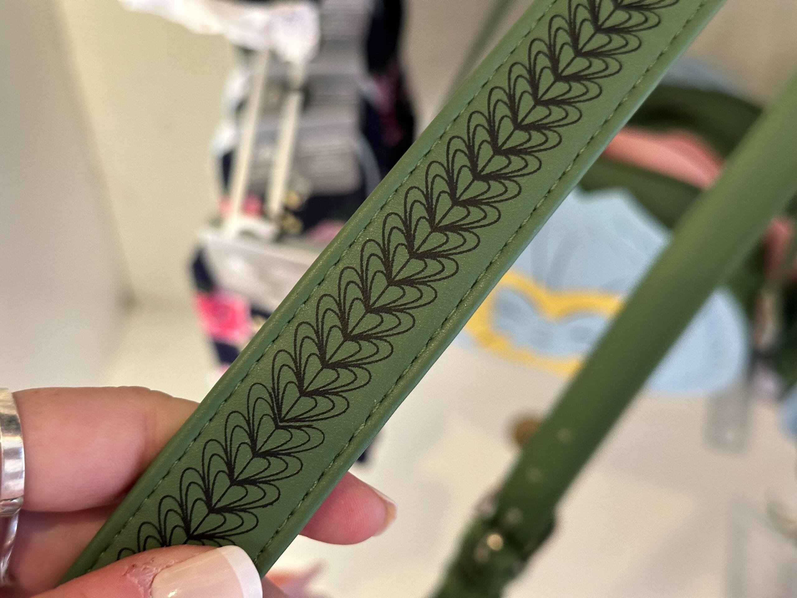 A close-up of a hand holding a green strap with a black, leaf-like pattern evokes the whimsical essence of "Alice in Wonderland." The background is tastefully blurred, highlighting clothing items that resonate with Ashley Eckstein's vibrant style.