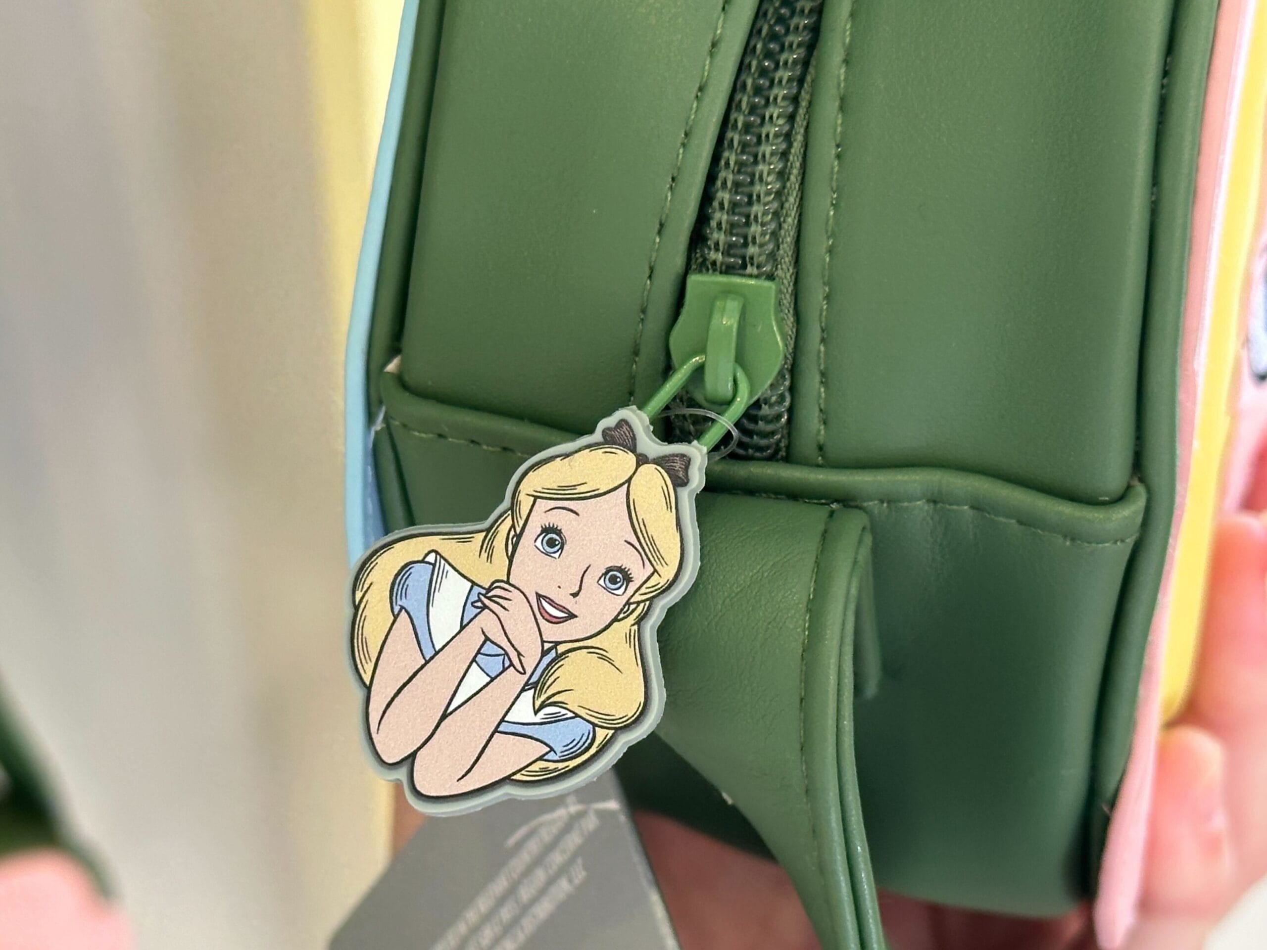 A green bag with a zipper pull showcasing an illustration of a blonde girl, reminiscent of Alice in Wonderland, with a blue headband, smiling with her hands under her chin.