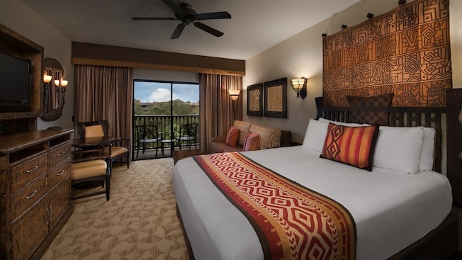 Hotel room with a large bed, patterned pillows, a carved wooden headboard, TV, wooden furniture, and a balcony view with outdoor chairs. Neutral tones and tribal-inspired decor.