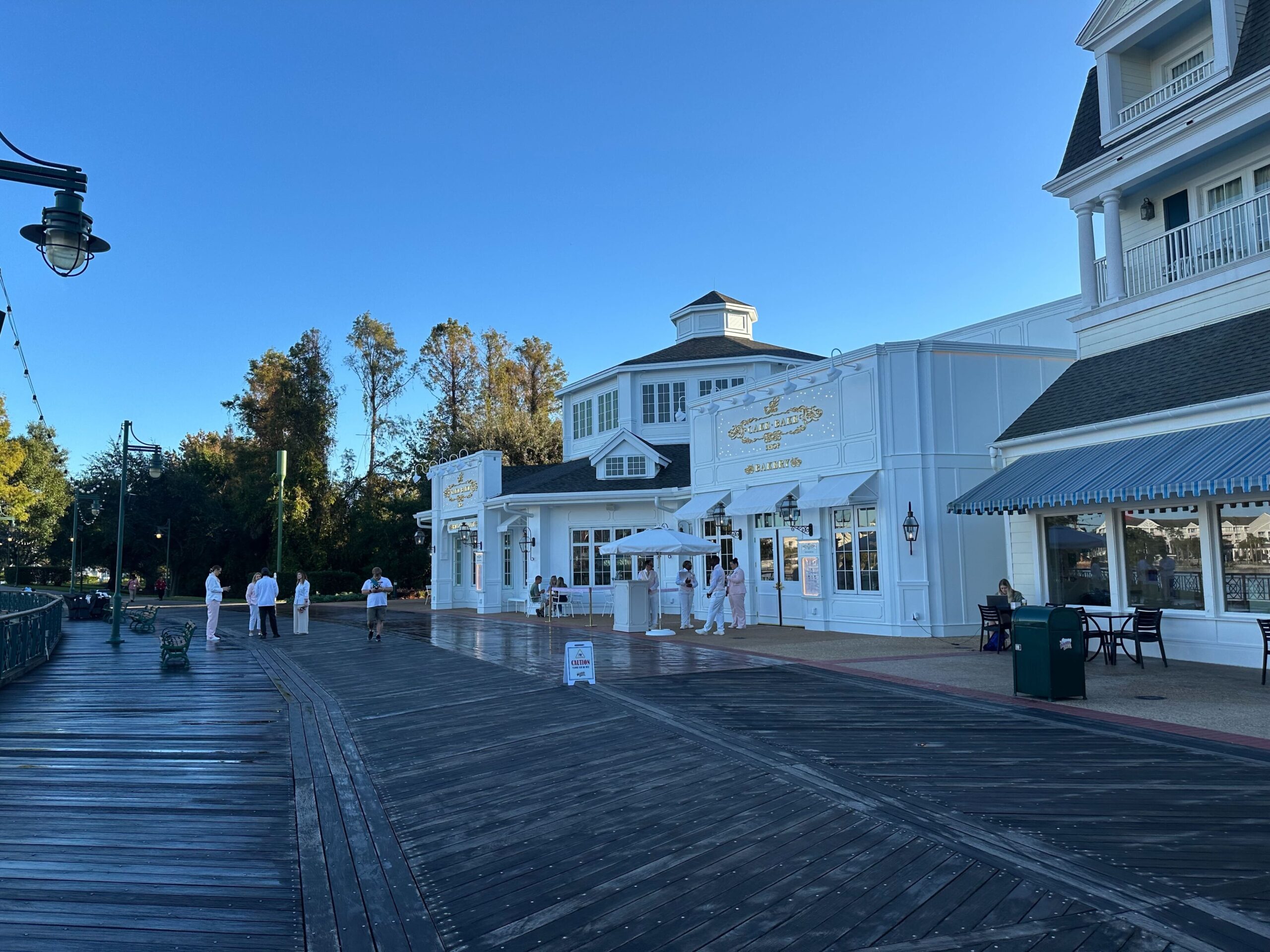 People stroll along a wooden boardwalk beside a white building with a sign that reads "BoardWalk Ice Cream." The scent of freshly baked treats from the nearby cake bake shop fills the air under the clear blue sky.