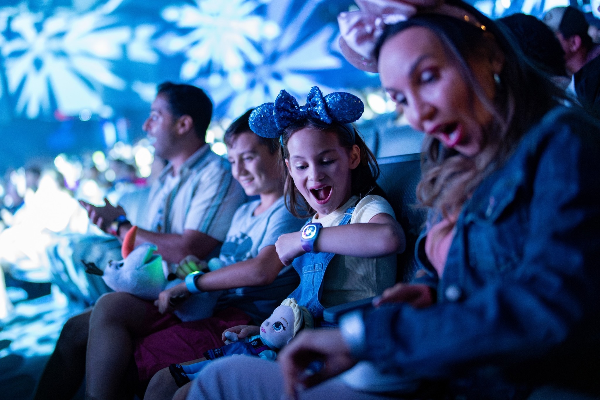 A family is sitting indoors, surrounded by blue lighting. A child wearing a bow headband looks excitedly at a wristwatch, while an adult next to them reacts happily.