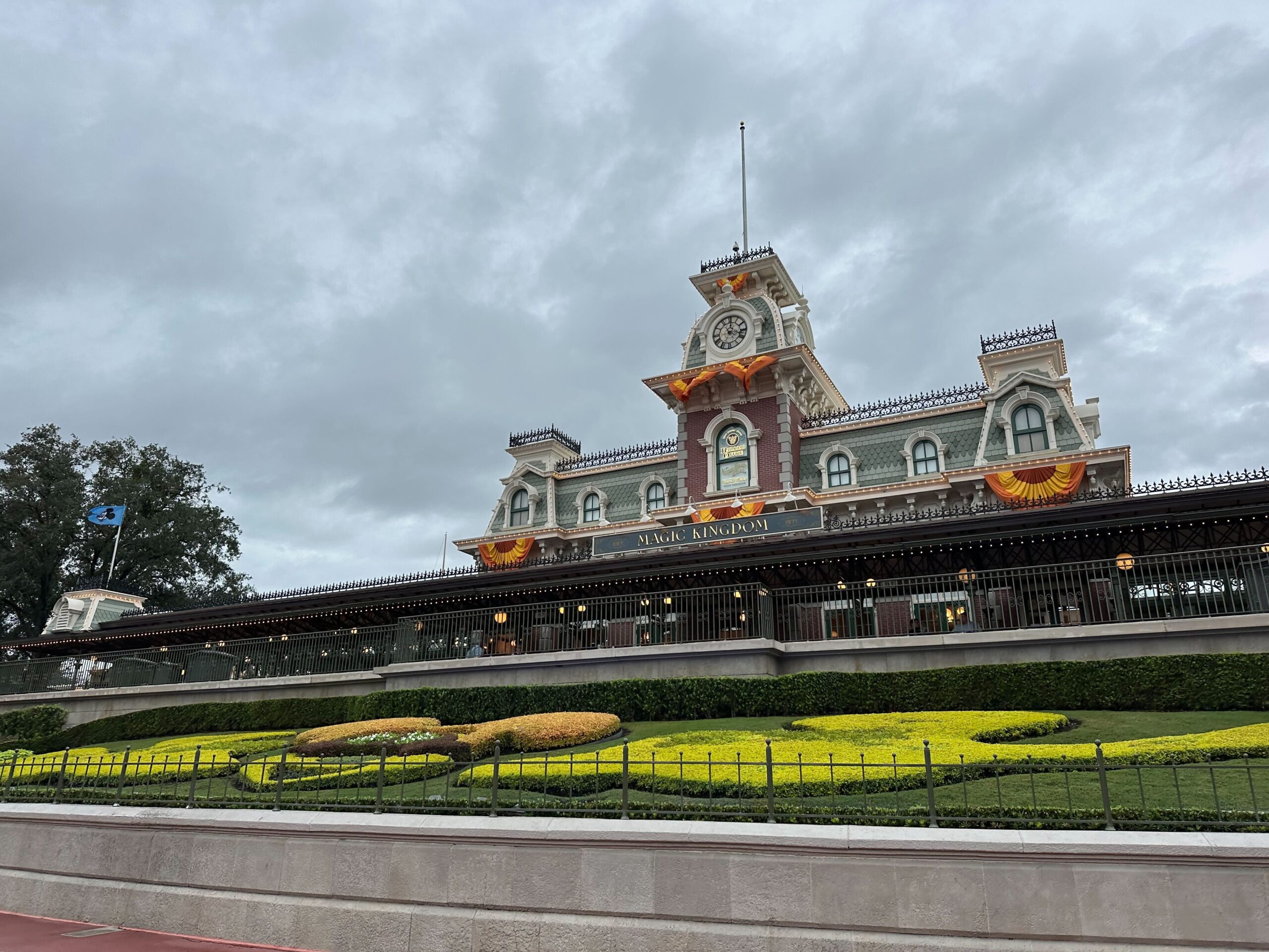 Train station with a clock tower and landscaped garden in front, under a cloudy sky.