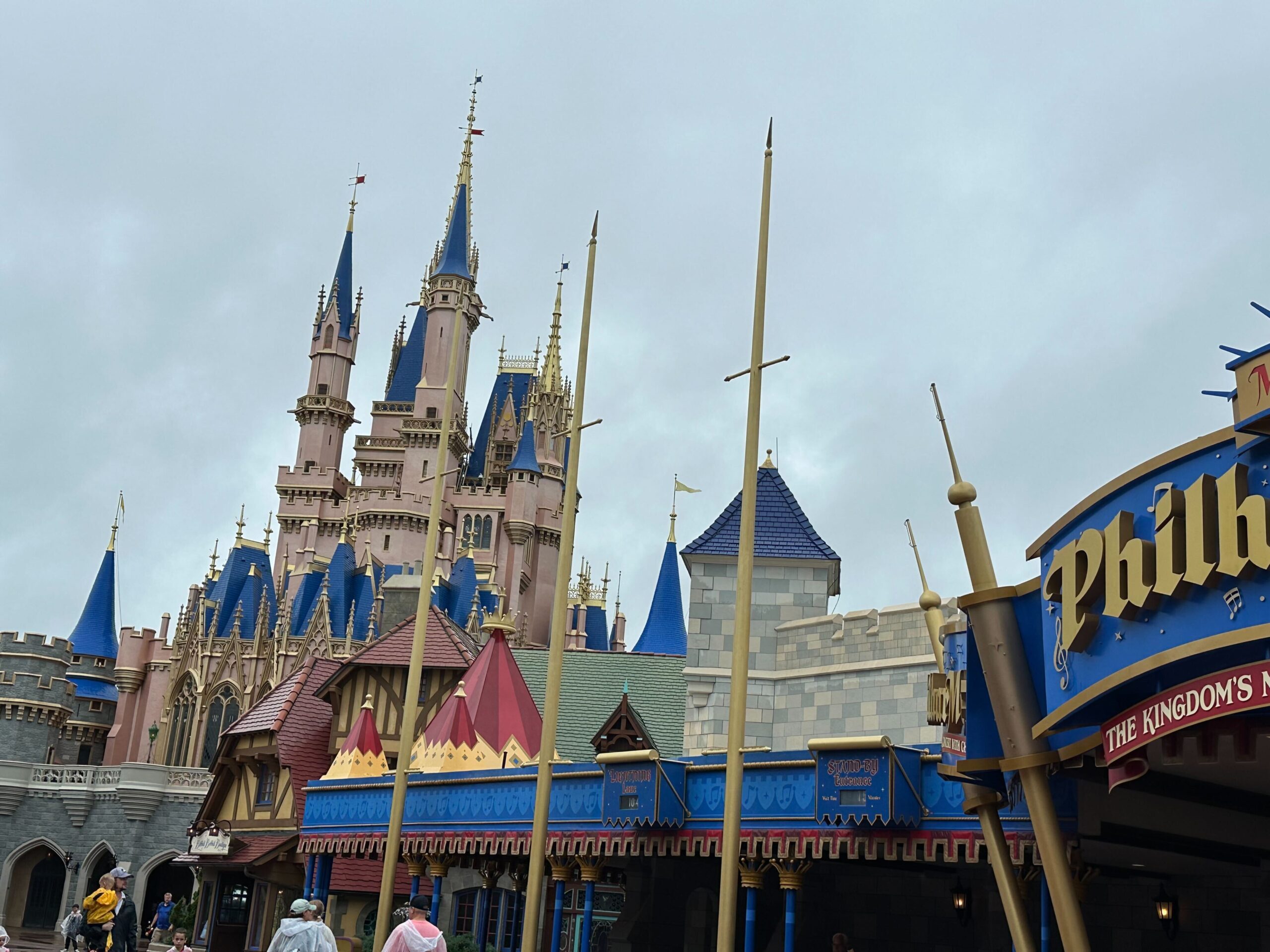 Amidst a cloudy sky, the fairytale castle with its blue spires stands majestically in the Magic Kingdom, surrounded by colorful buildings. Despite distant memories of Hurricane Milton, people stroll nearby, enchanted by the whimsical atmosphere.