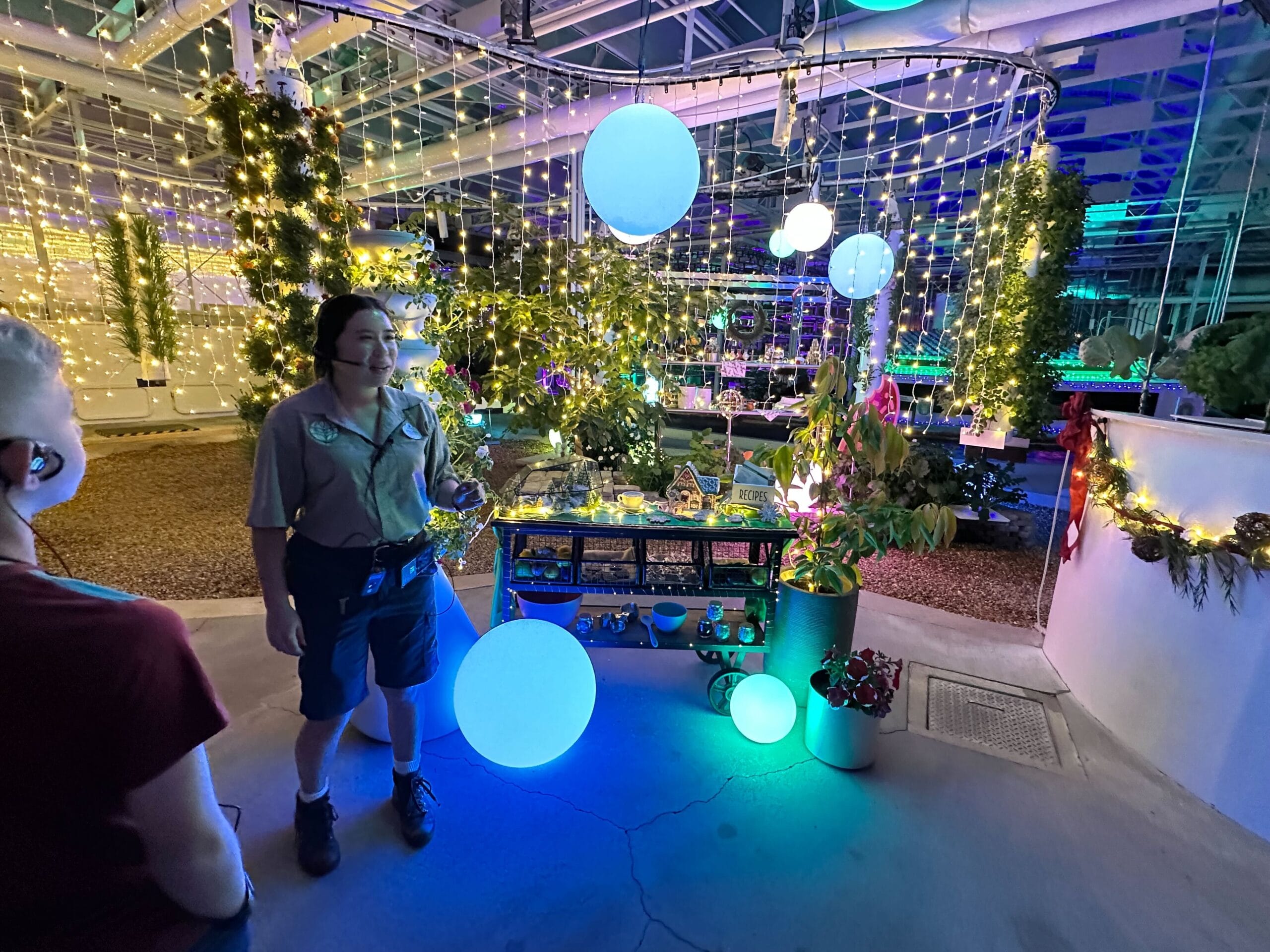 A person stands beside a lit display table in a glimmering greenhouse adorned with lights and plants, as part of the enchanting Behind the Seeds Tour. Another individual is partially visible in the foreground, adding to the immersive Living with the Land experience.