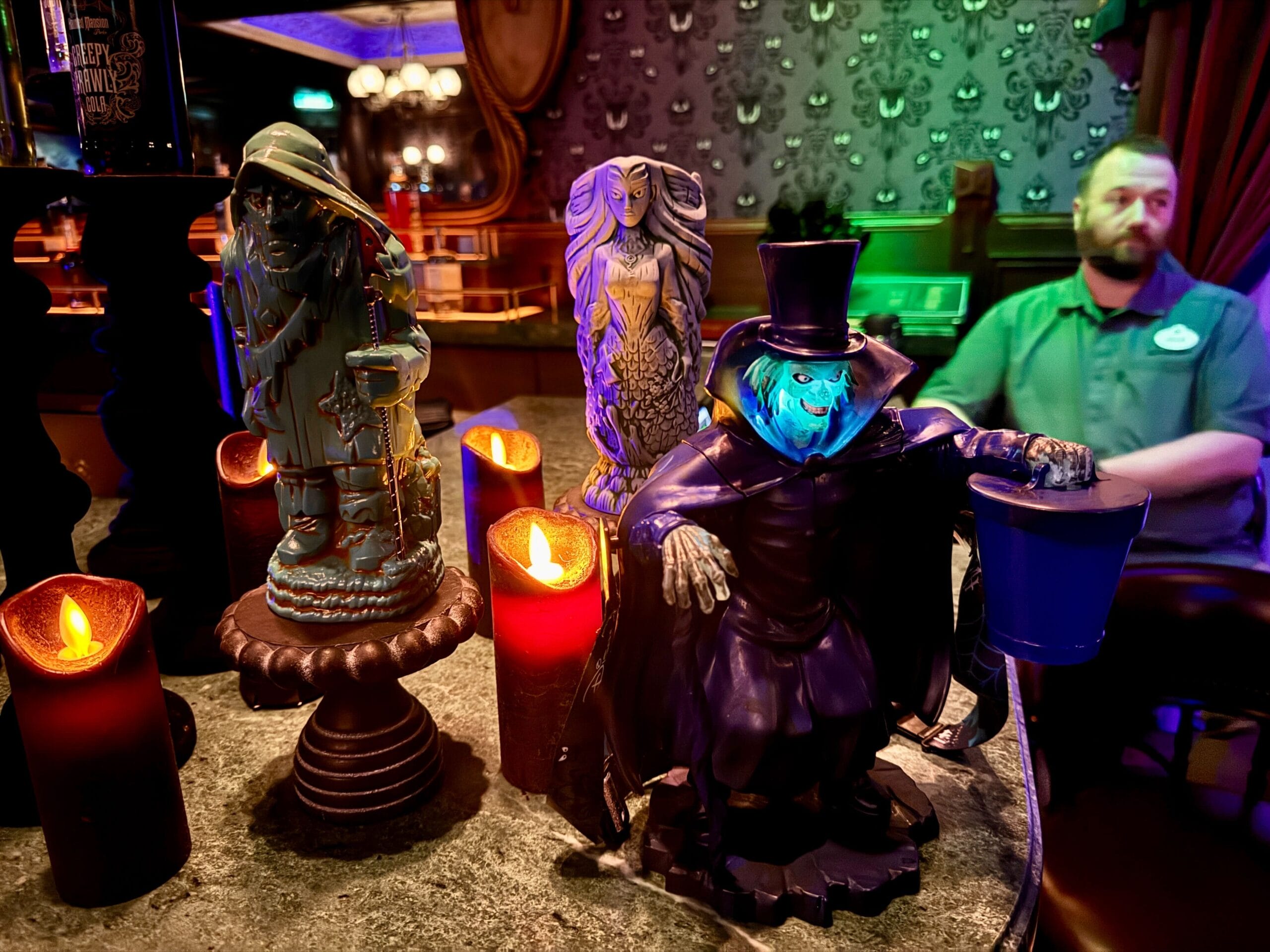 Three eerie statues are surrounded by glowing candles on a table in a dimly lit room. A person is seated in the background.
