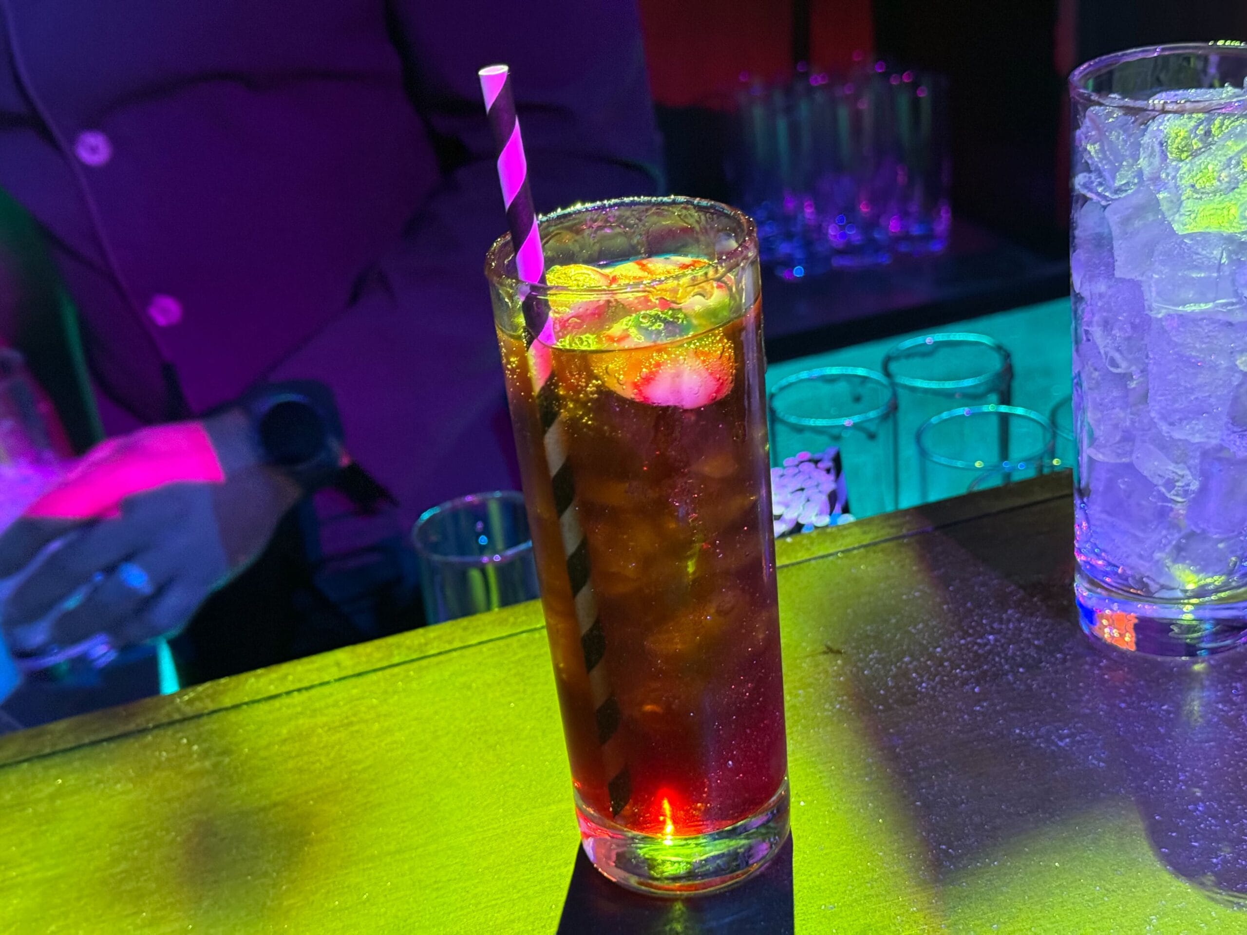 A tall glass of cola with ice and a striped straw on a bar counter, surrounded by colorful lighting.