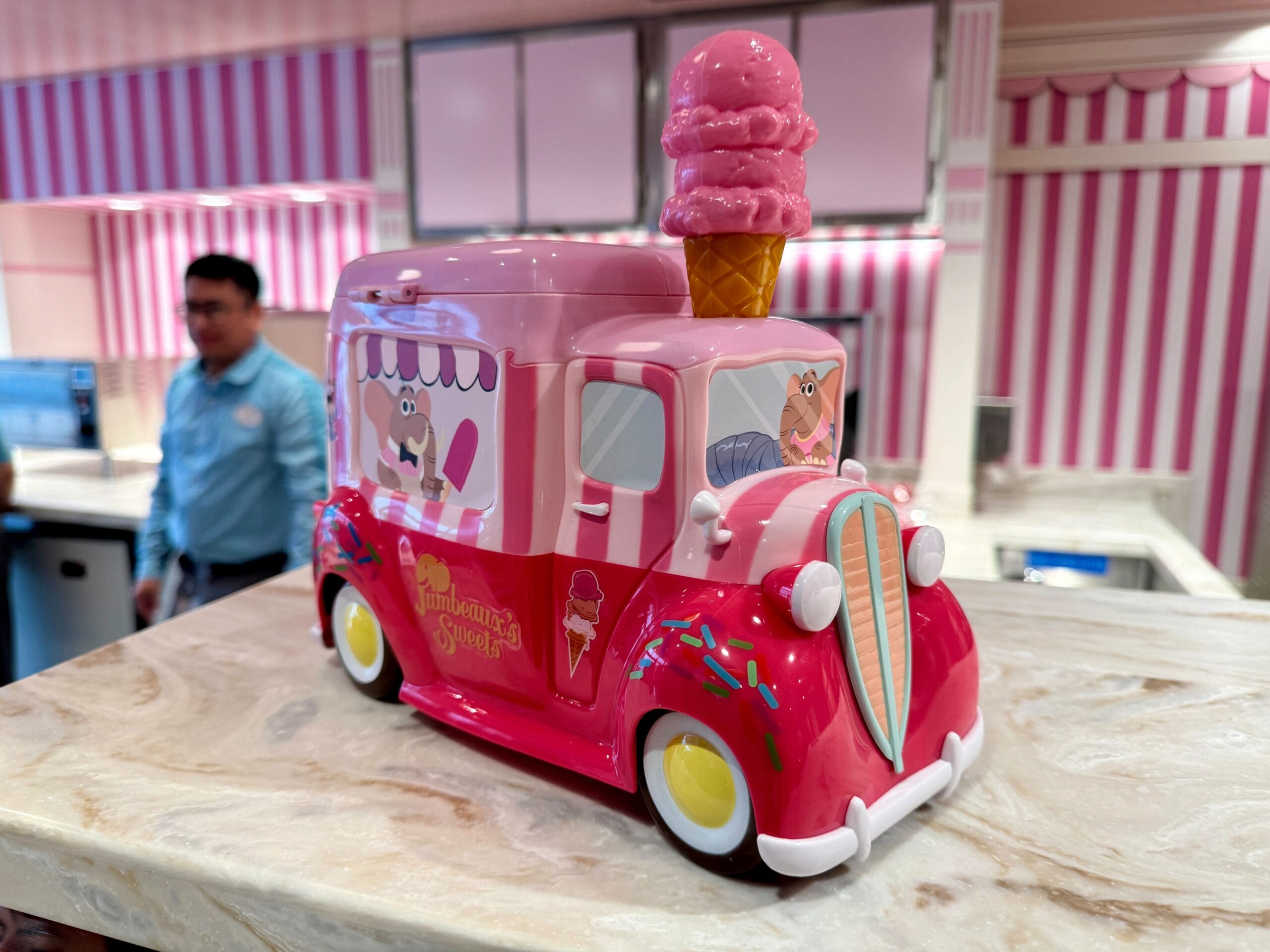 A pink toy ice cream truck with an ice cream cone on top sits on a counter. A person in a blue shirt stands in the background.