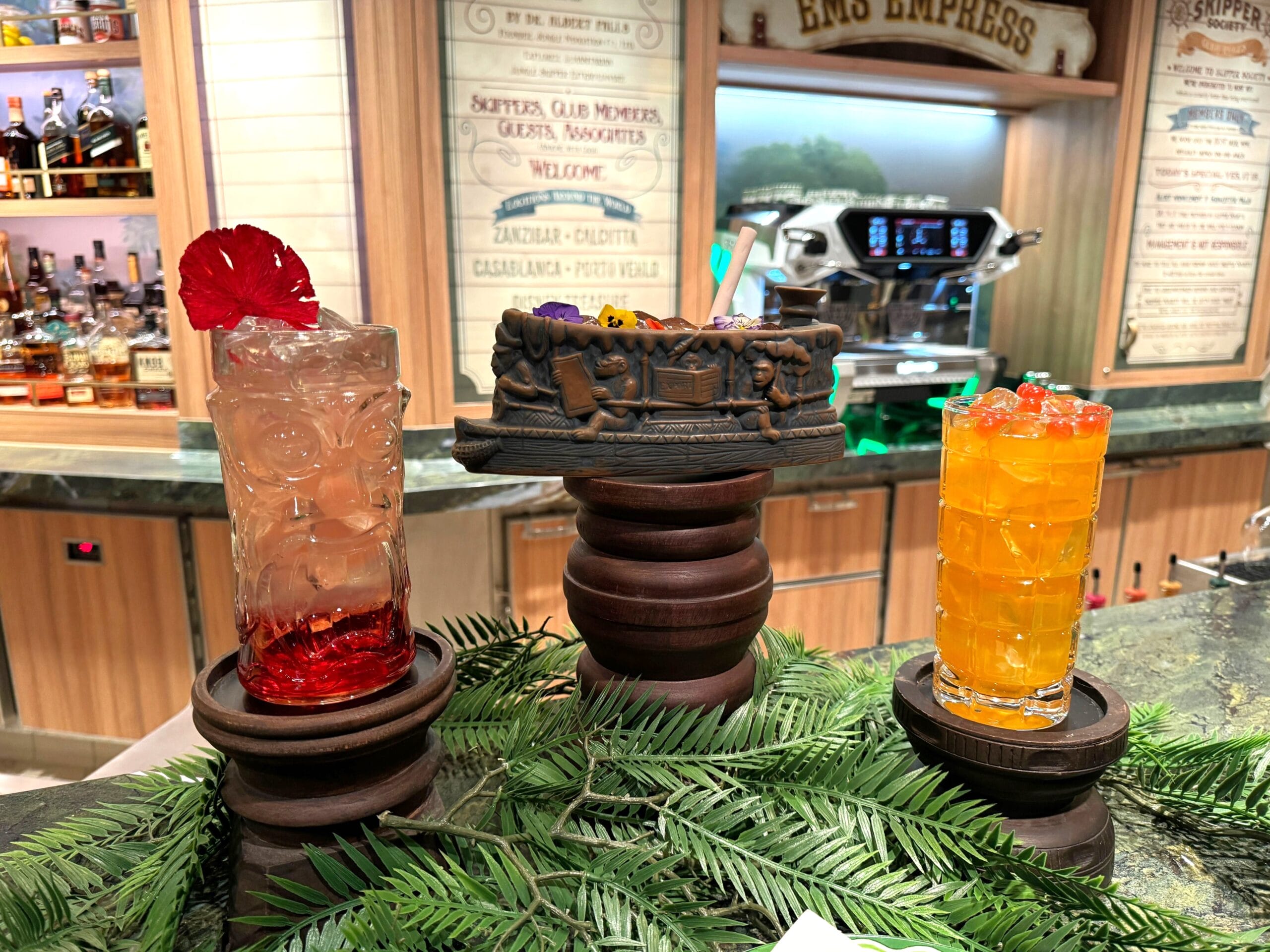 Three cocktails on wooden pedestals: one with a red flower, one in a decorative mug, and one with orange liquid. Background features a bar and a coffee machine.