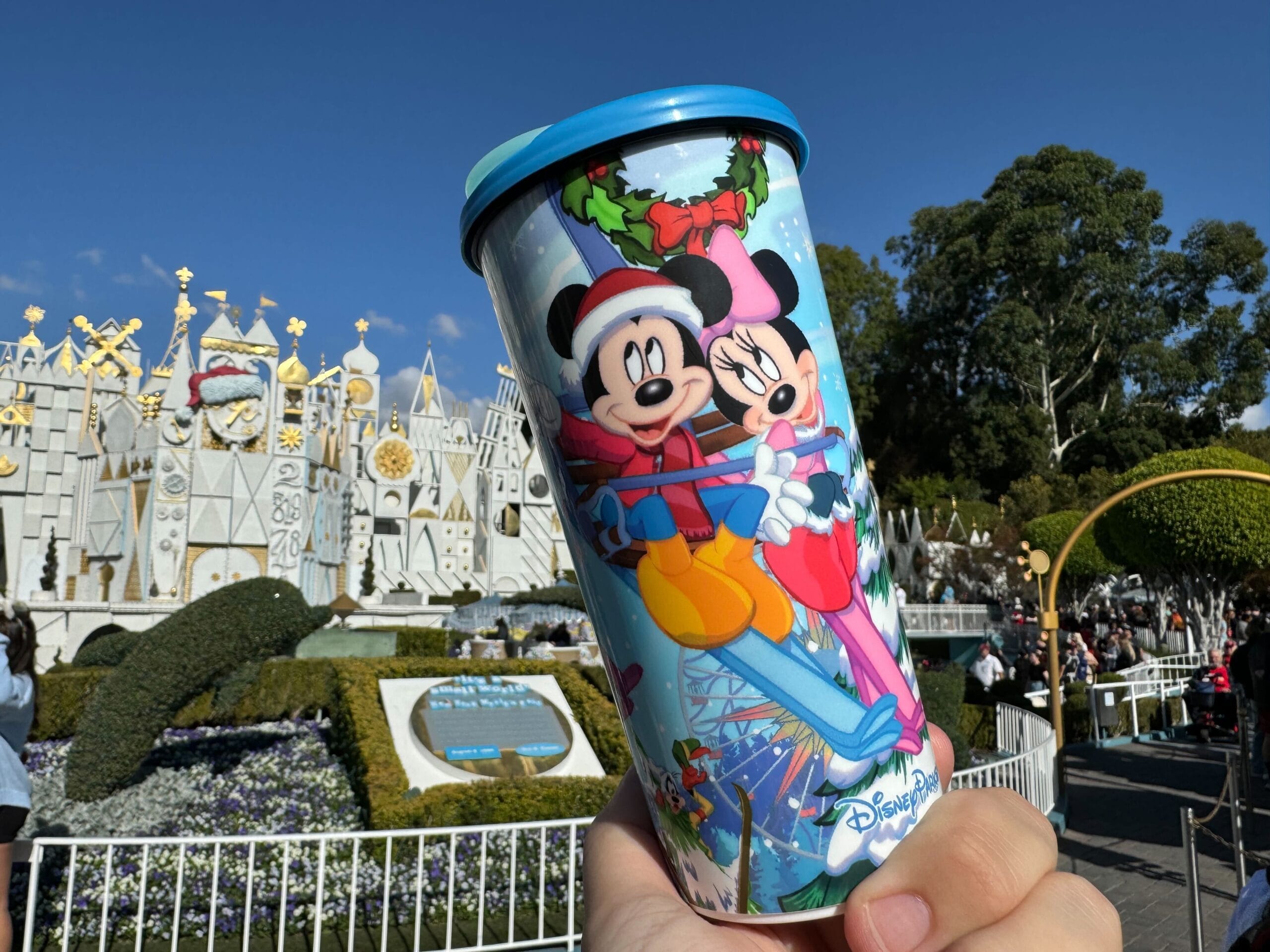 A hand holds a vibrant Disney-themed 2024 holiday travel tumbler adorned with Mickey and Minnie Mouse, perfectly capturing the charm of the "It's a Small World" attraction at the theme park.