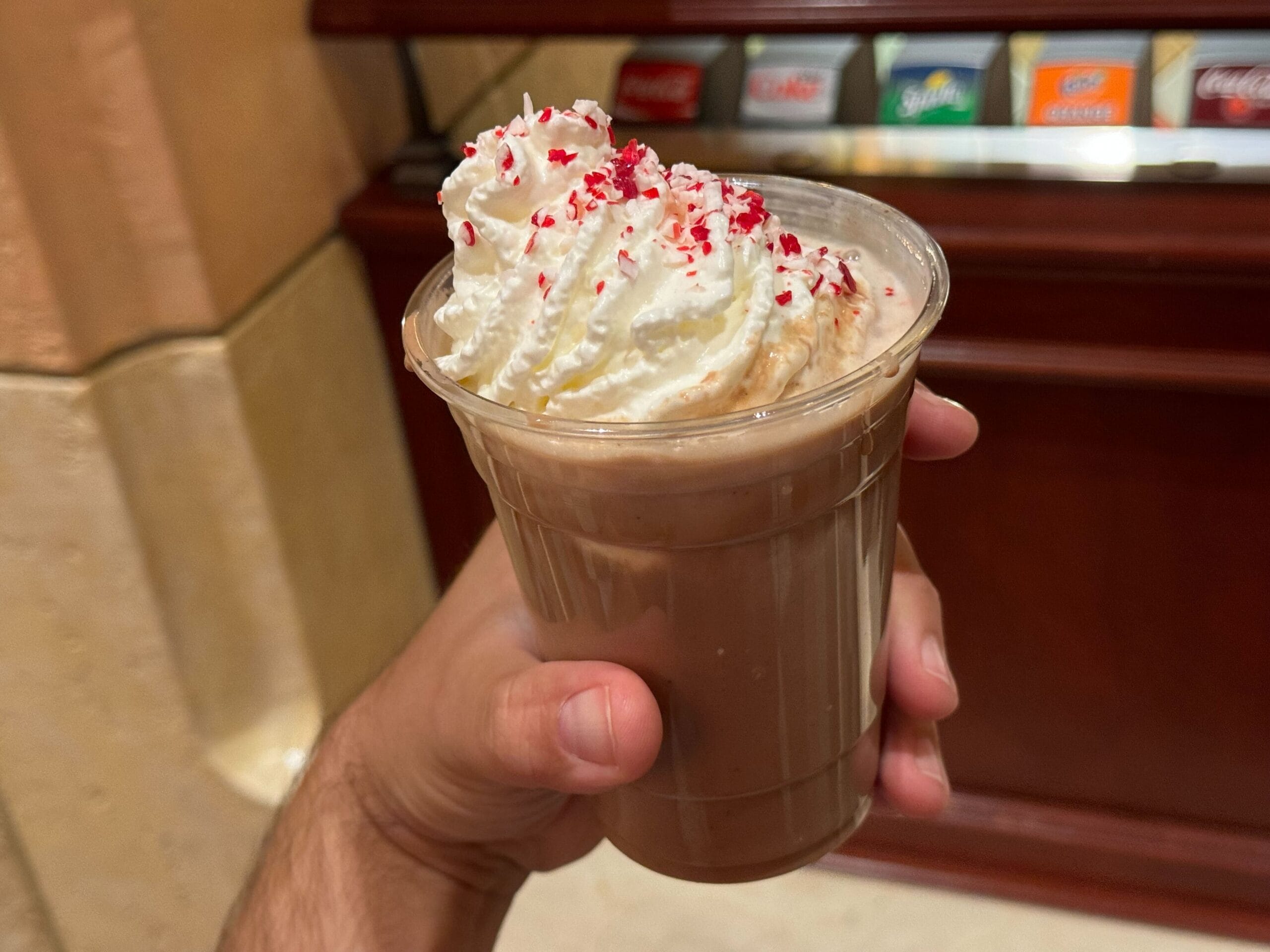 A hand holds a clear plastic cup of Warm Chocolate Float, topped with whipped cream and festive red sprinkles, perfect for the 2024 Holidays. In the background, a wooden counter is nestled near soda dispensers at Disney California Adventure.