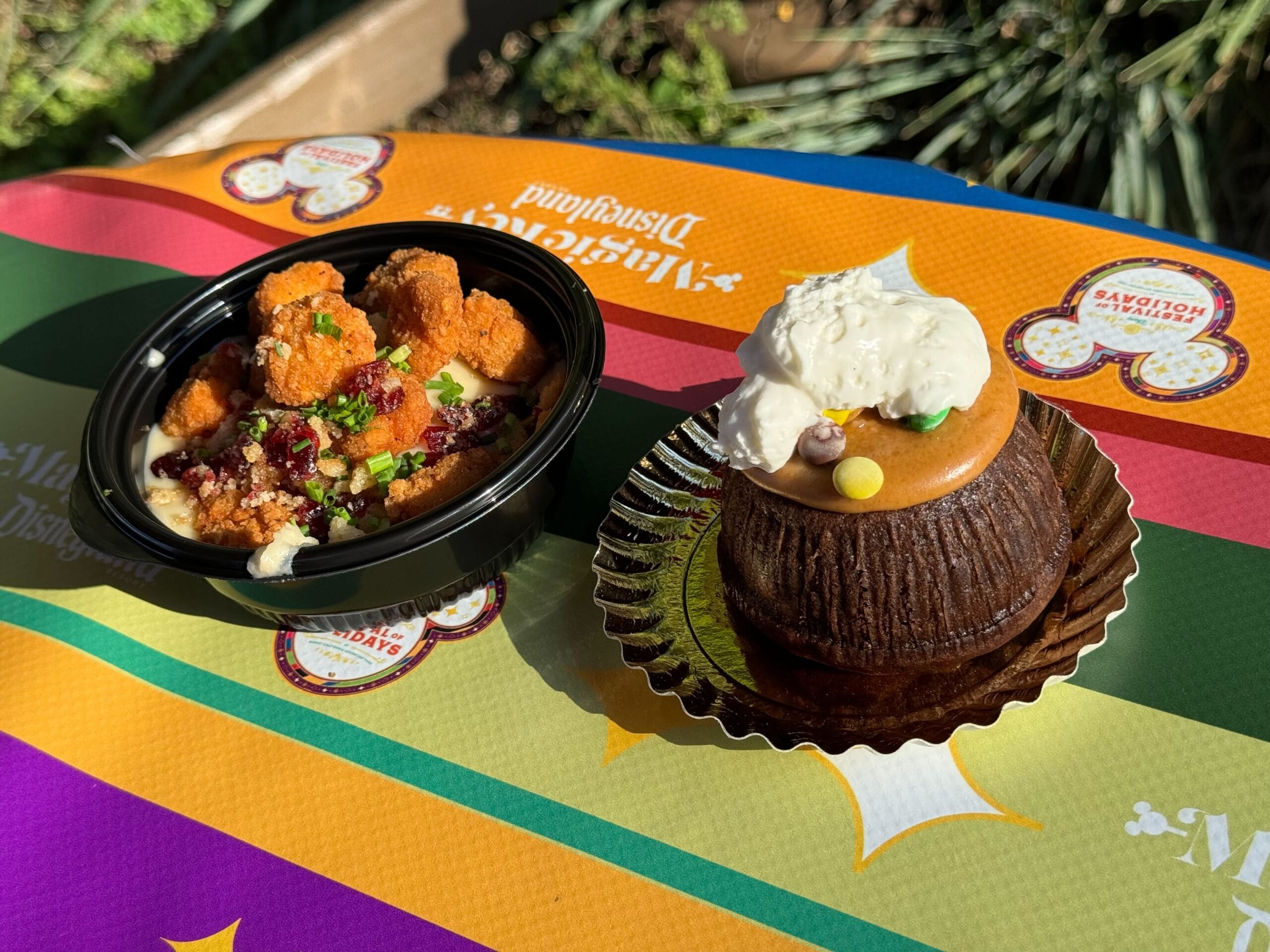 A black bowl with fried chicken pieces, garnish, and sauce sits next to a chocolate cupcake adorned with orange frosting and whipped cream on a colorful tablecloth—an inviting scene of Merry Mashups perfect for any festive feast.